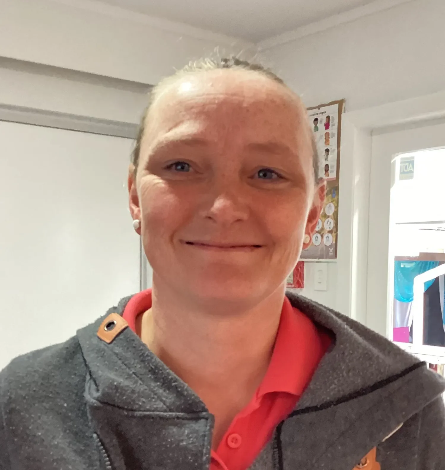 A woman smiling, wearing a grey jacket with a brown button detail, and a red shirt underneath, standing indoors.