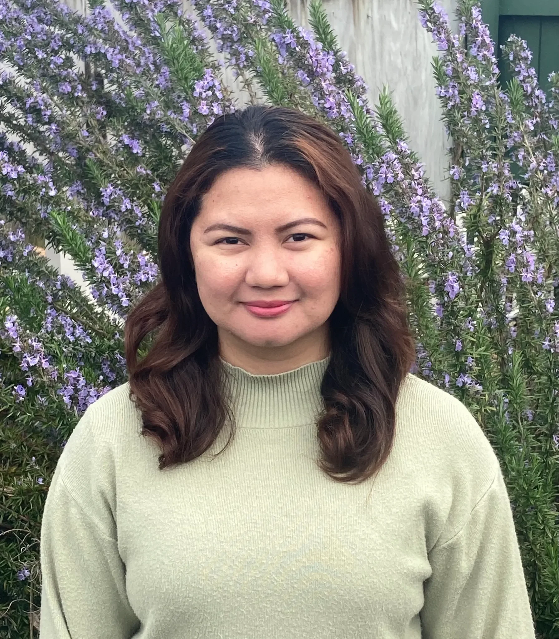 A woman with shoulder-length brown hair smiles softly at the camera, standing outdoors in front of a large bush with purple flowers.
