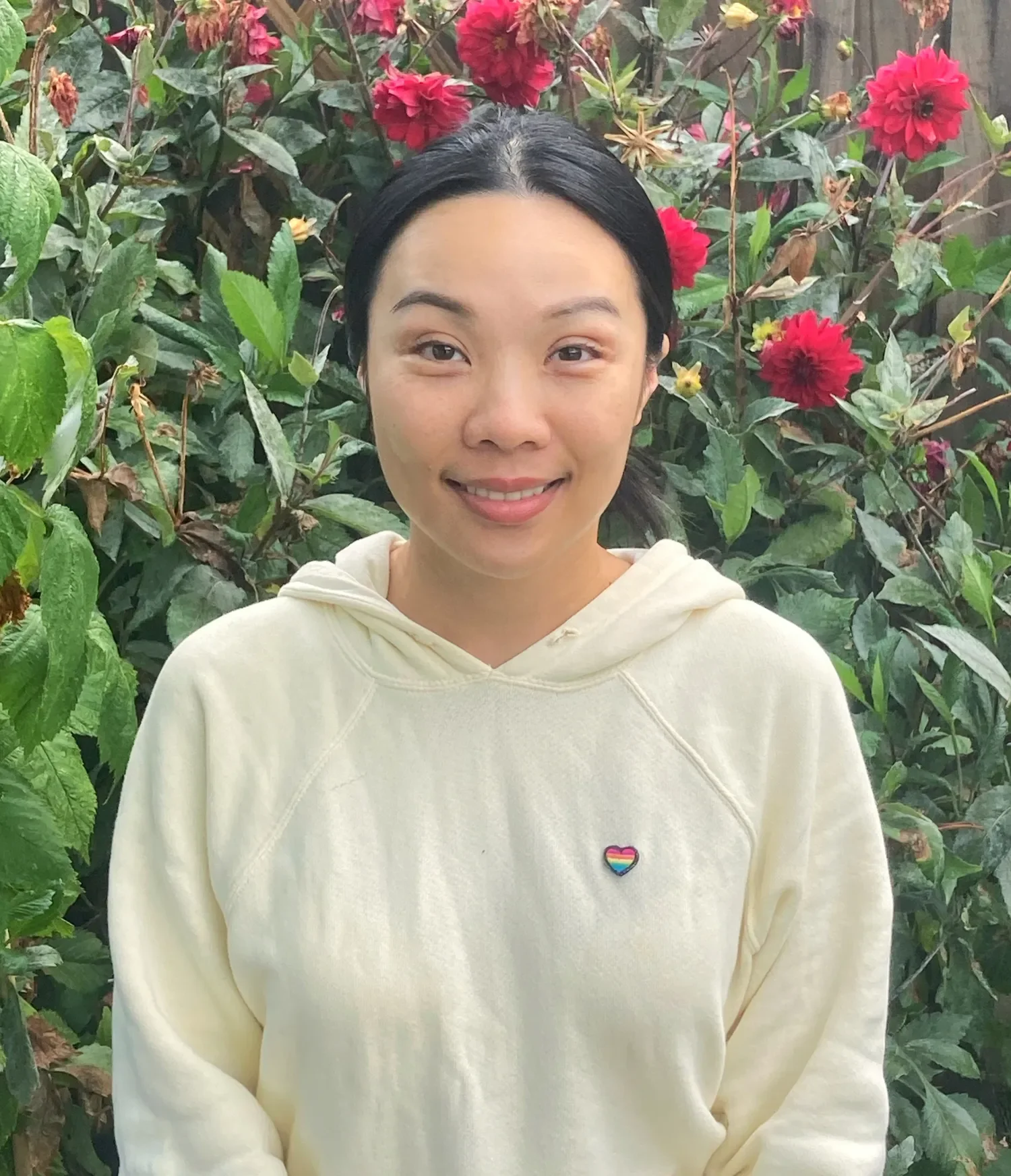 A woman with black hair wearing a cream-colored hoodie standing in front of a bush with red flowers and green leaves, smiling at the camera.