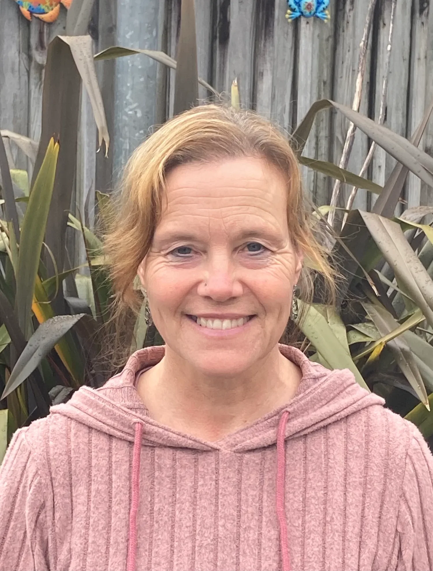 A woman with short, wavy red hair and blue eyes smiling outdoors in front of a wooden fence and plants.