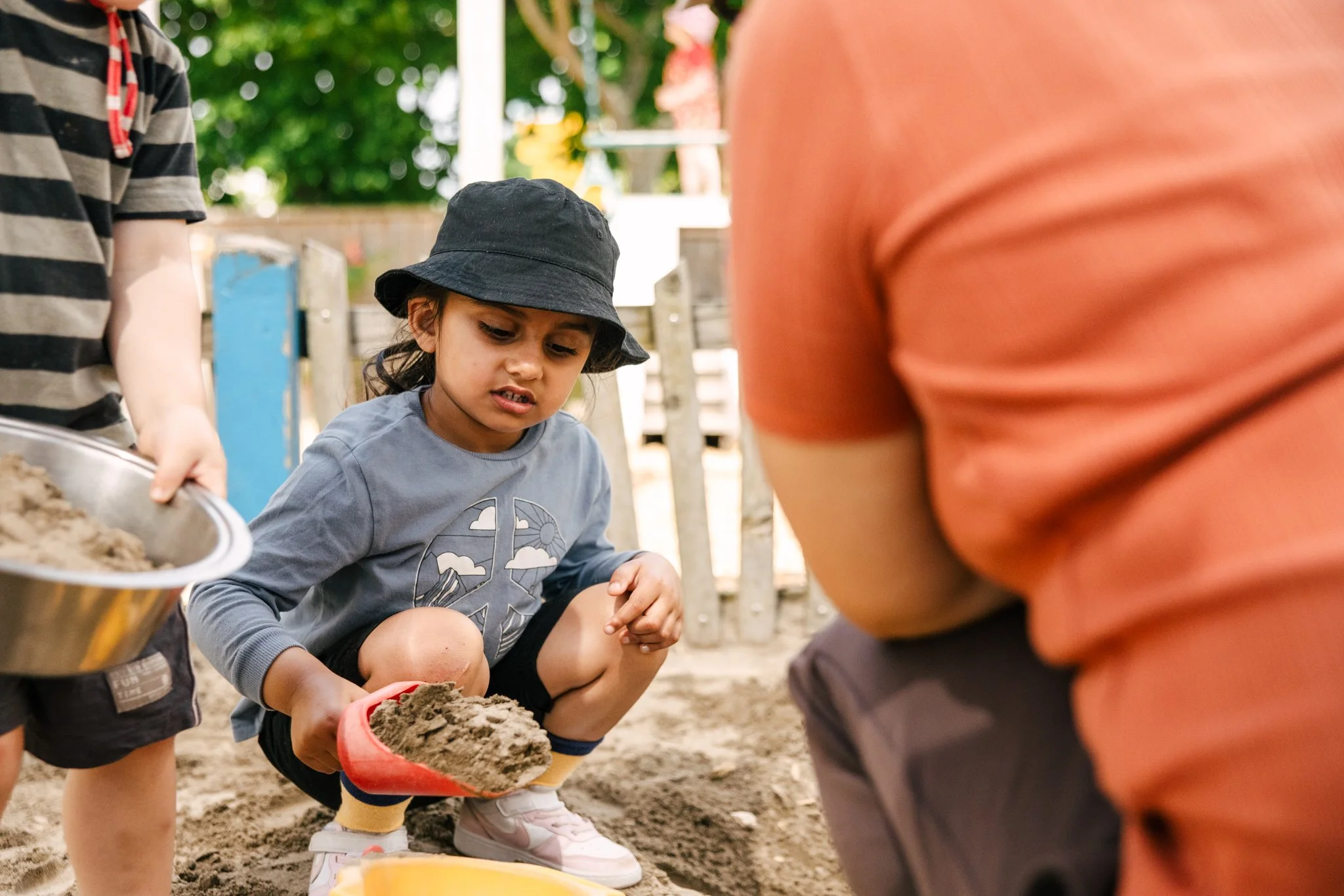 A young girl wearing a black hat and gray shirt playing in the sandbox with a red shovel, while other children hold tools nearby.