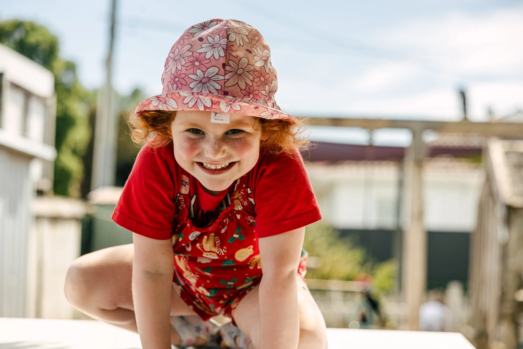Young girl with curly red hair smiling and crawling outdoors, wearing a pink floral hat and a red shirt, on a bright sunny day.