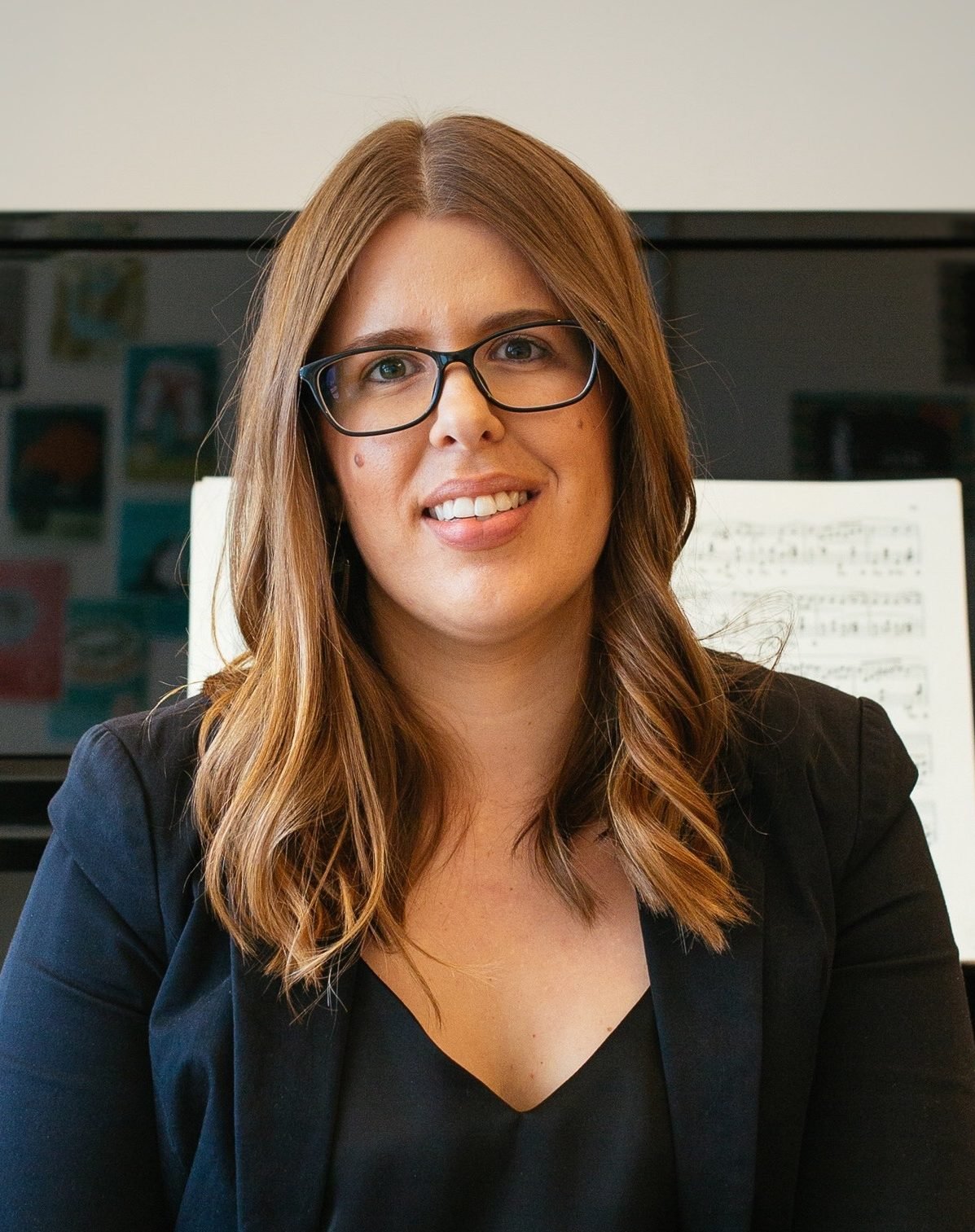 A woman with glasses and shoulder-length brown hair, smiling, wearing a black blazer and top, sitting in front of a music stand with sheet music.