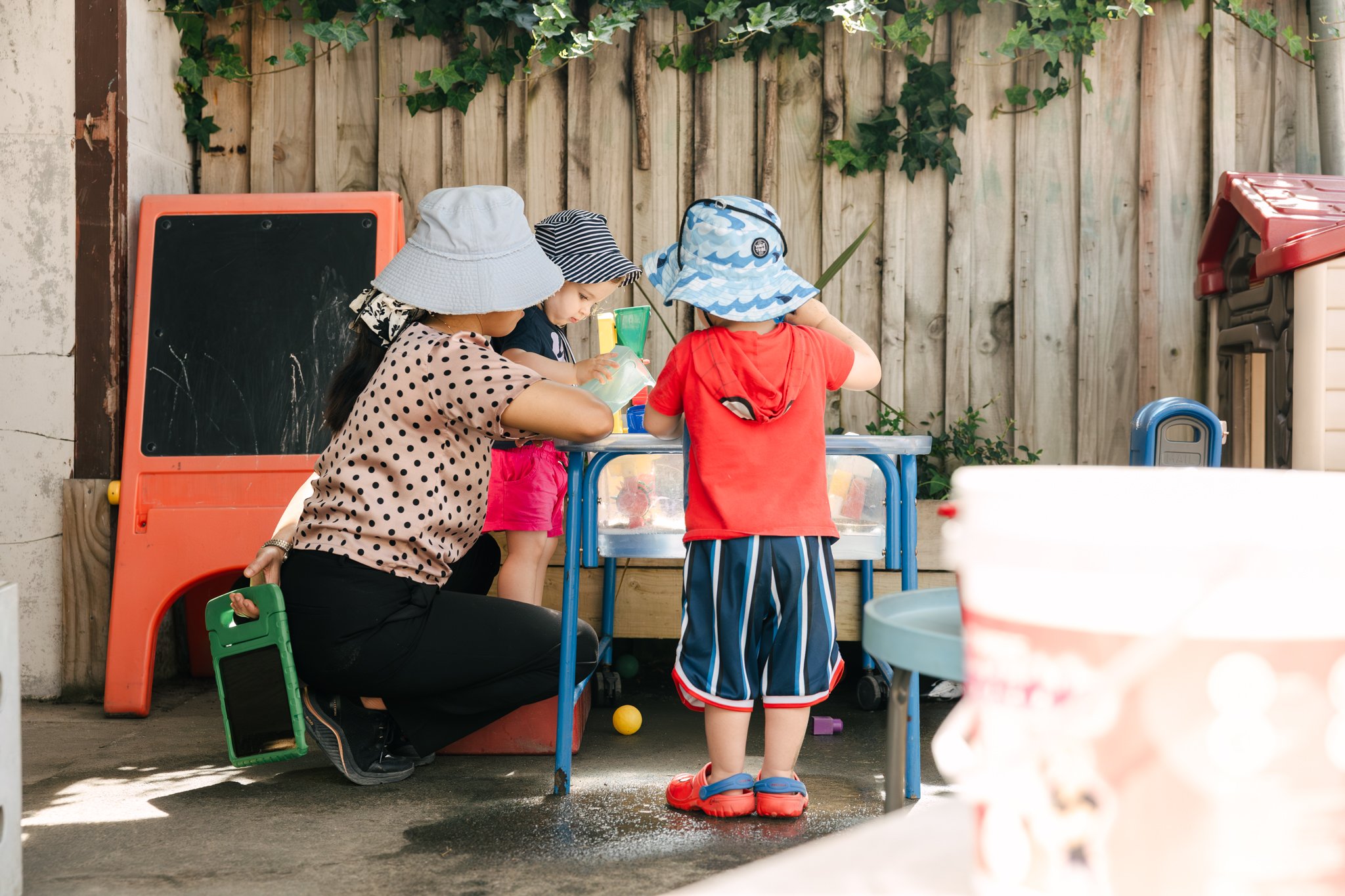 A woman with three children playing in a backyard. The woman is kneeling down while the children stand around a blue play table. They are all wearing hats, and the scene is set against a wooden fence with green vines.