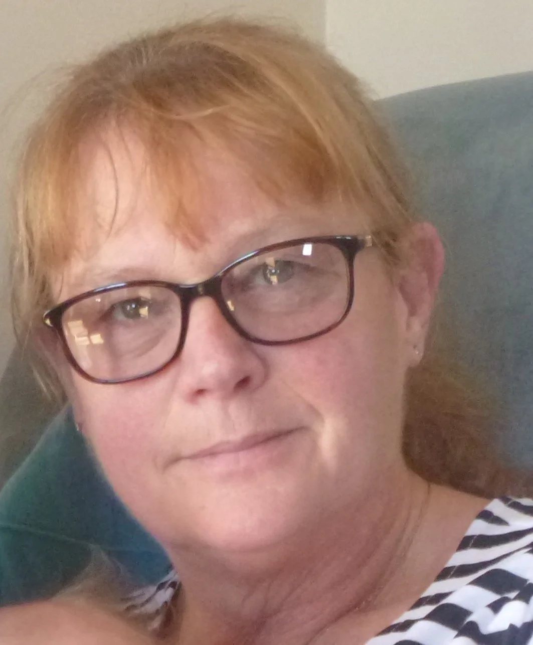 Close-up of a woman with light skin, red hair, glasses, and a black and white striped shirt, sitting in a chair.