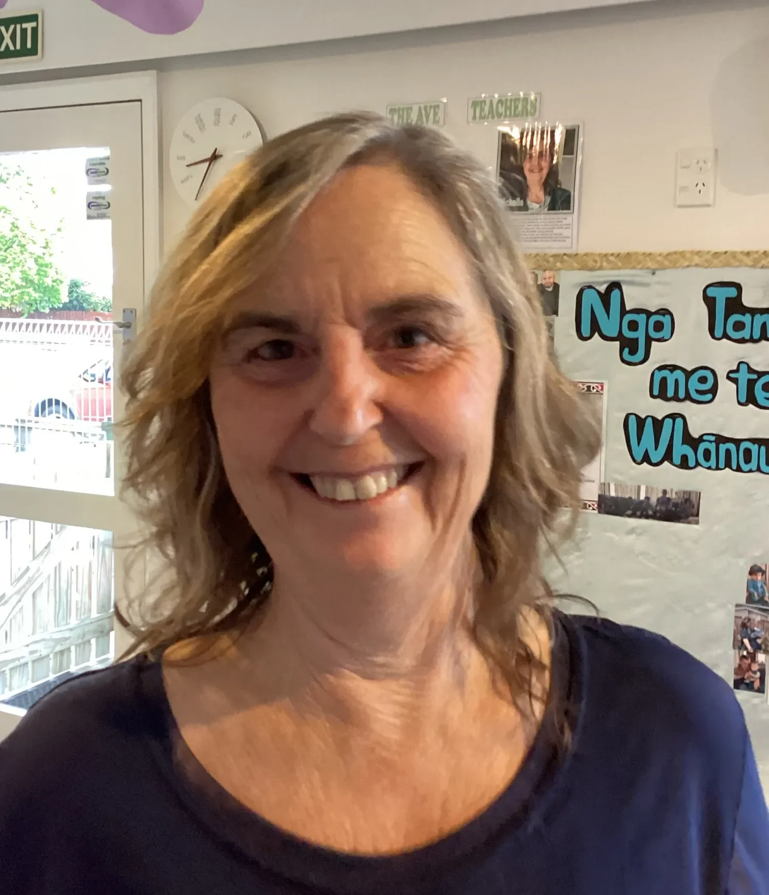 Smiling woman with wavy blonde hair indoors, behind her a bulletin board with blue and black writing and photos, and a white door with a window.