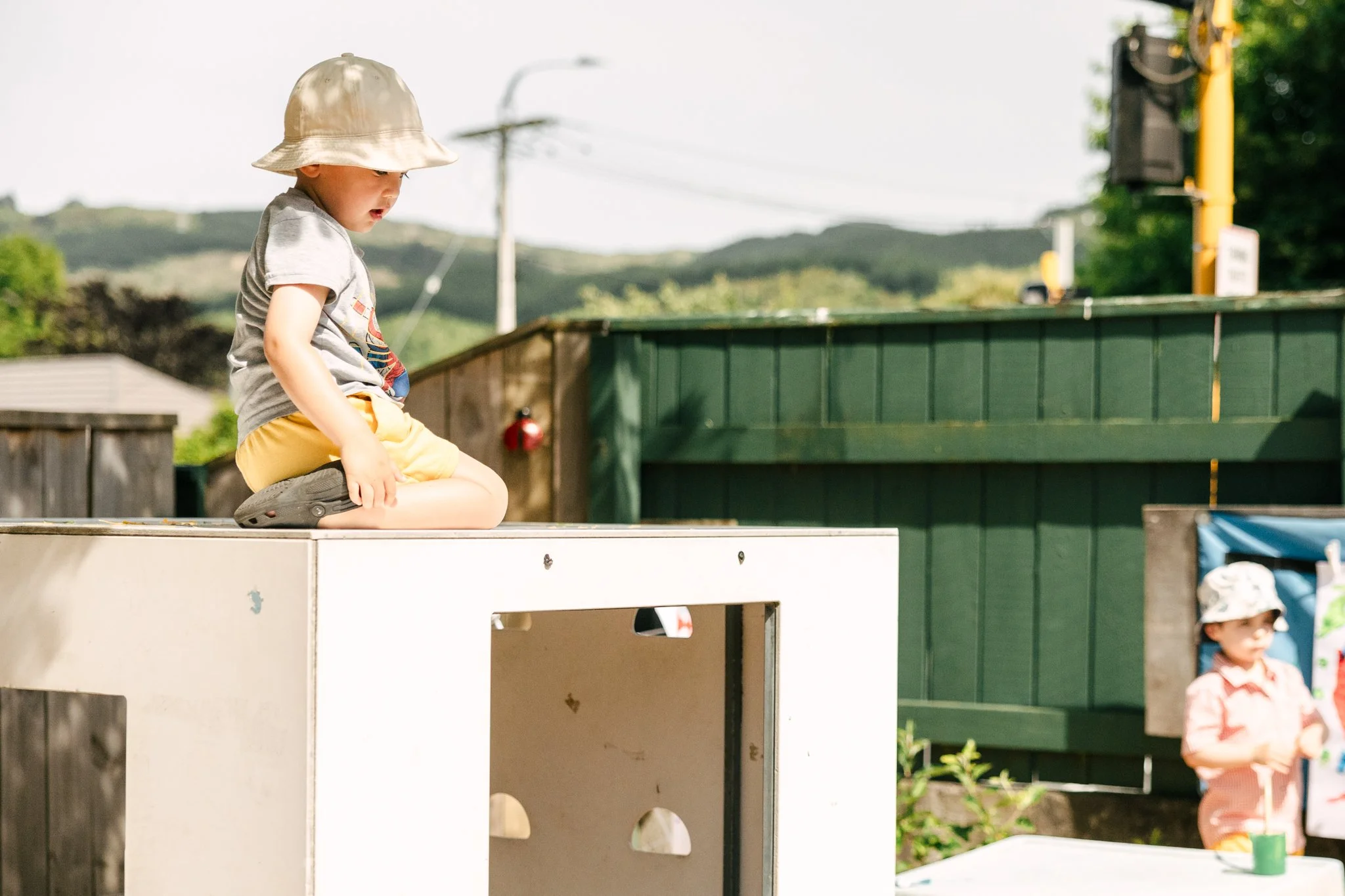 A young child wearing a beige hat, gray shirt, and yellow shorts sitting on a white playground structure outdoors.