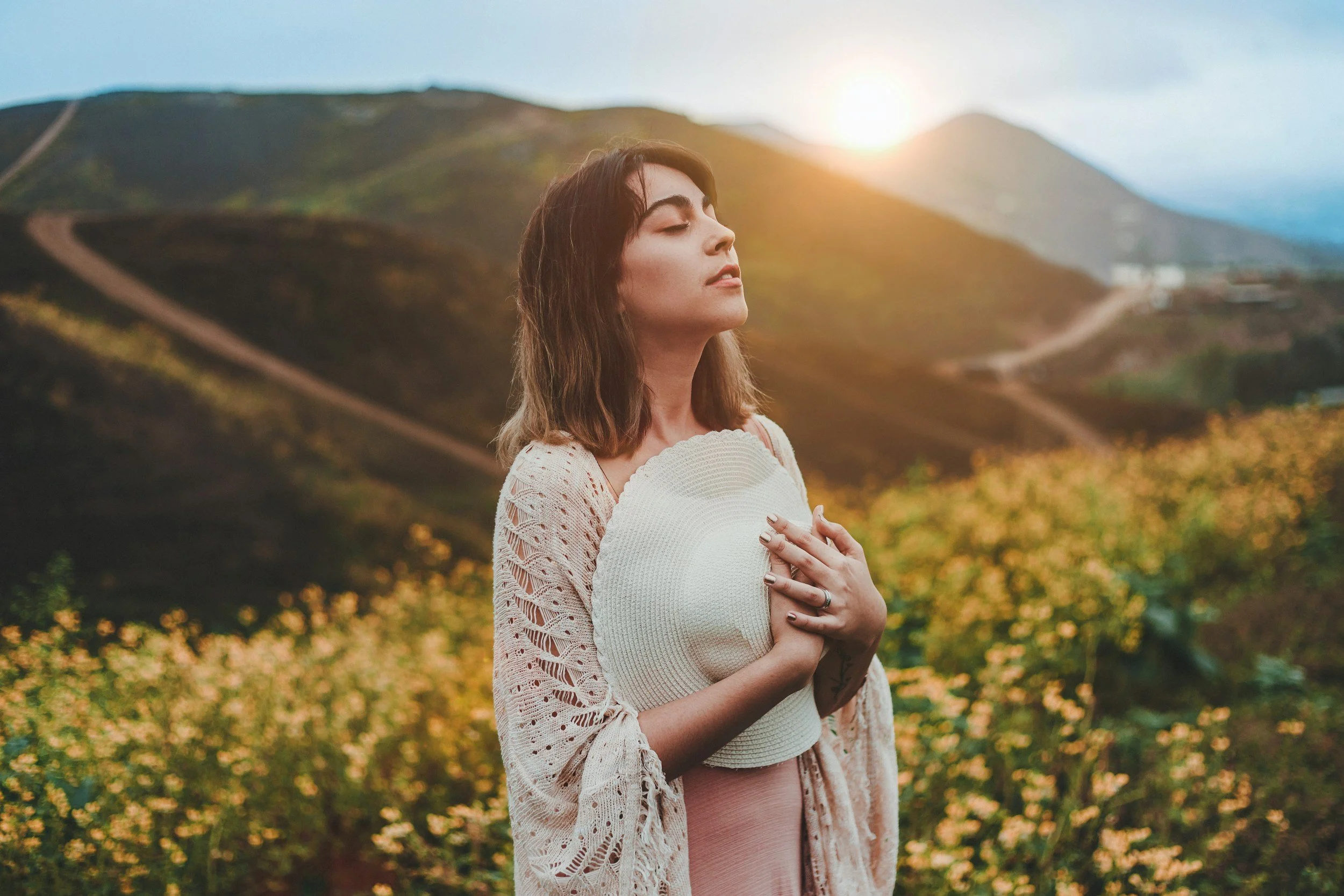 A young woman holding her heart with eyes closed, standing in front of softly lit mountains, evoking introspection, self-compassion, and emotional healing