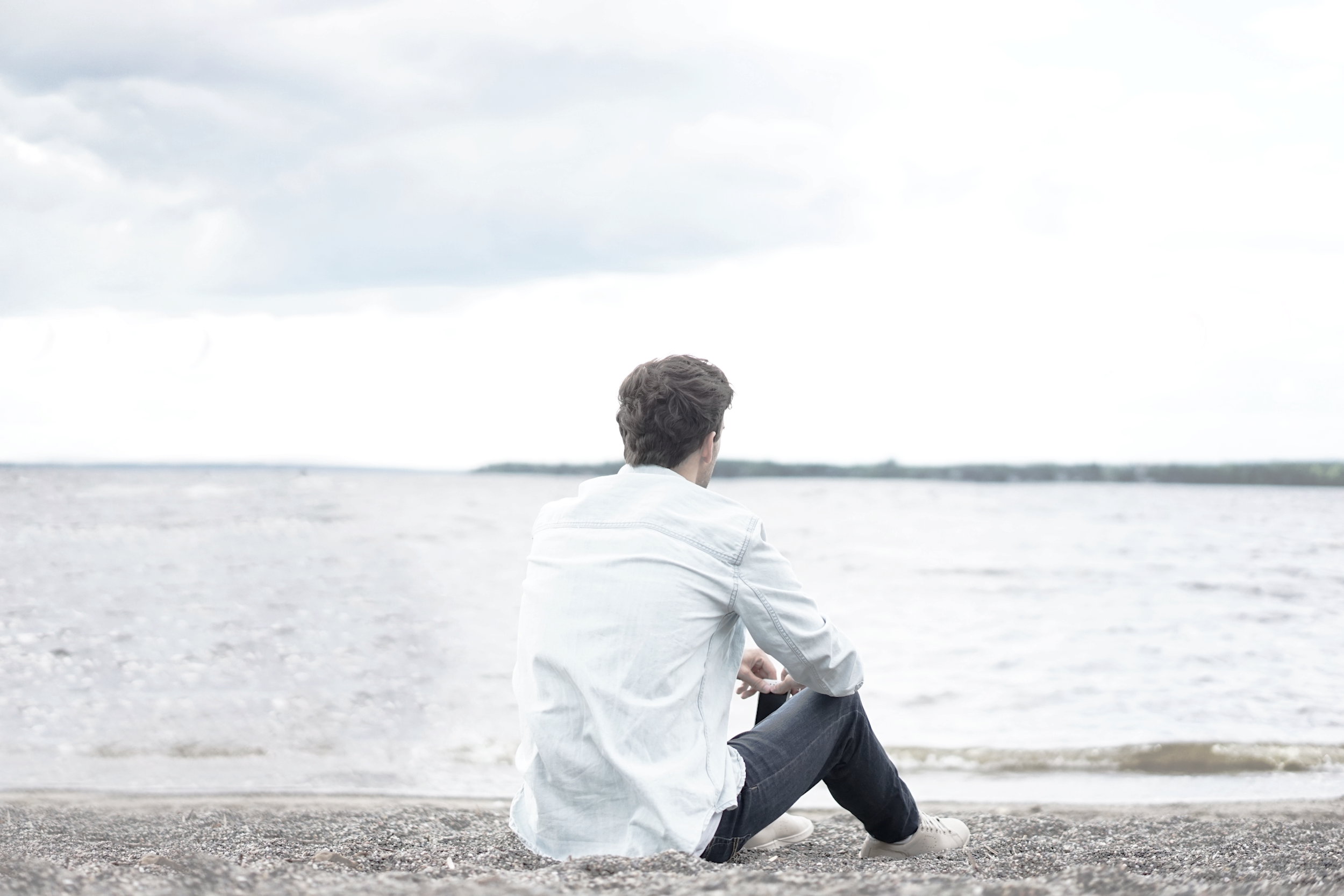 A man sitting by the water with his back to the viewer, looking toward open waters and a distant tree line, evoking reflection, calm, and contemplation
