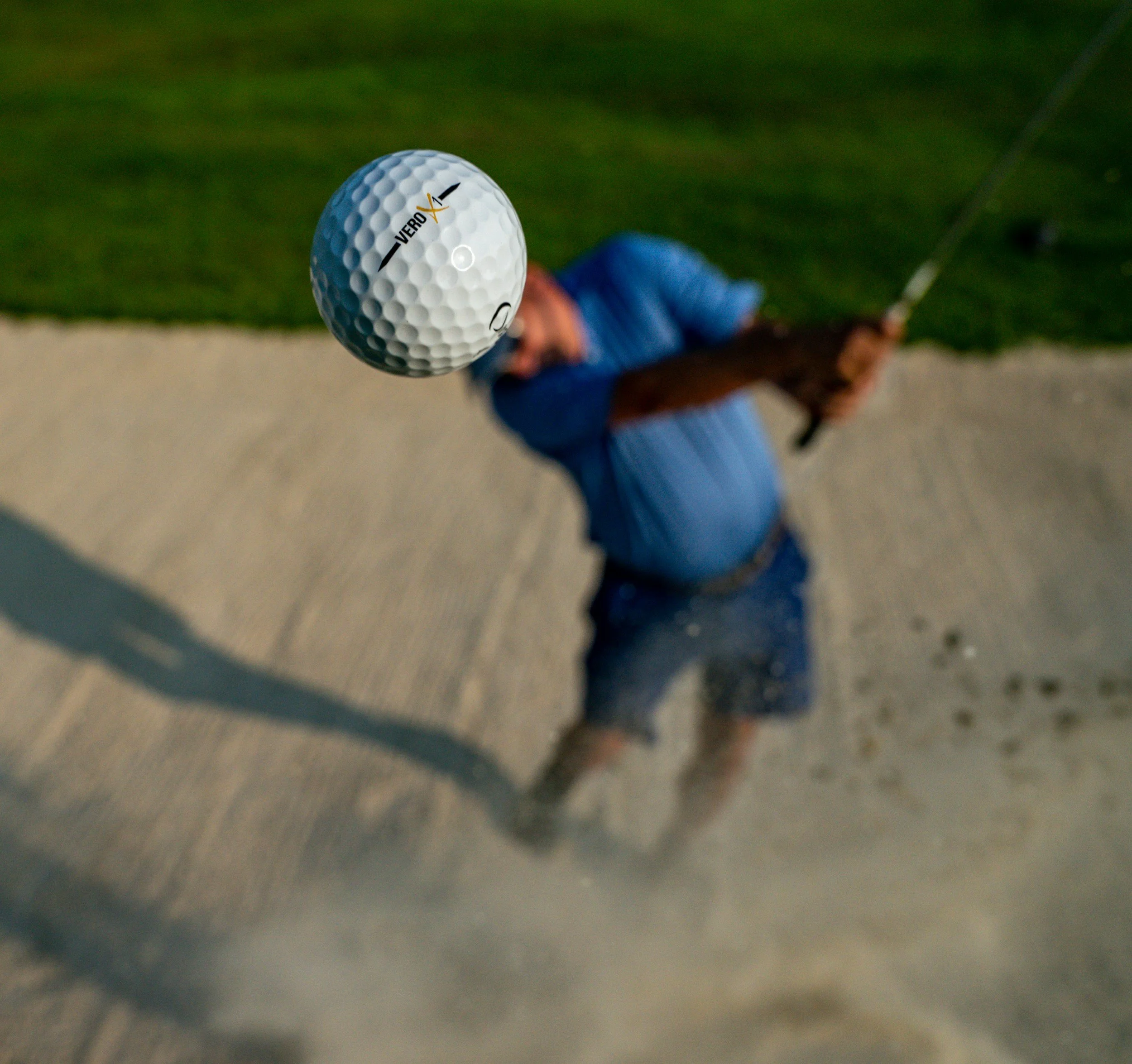 A person in a blue shirt swinging a golf club, hitting a golf ball with a "VeroX" logo, on a golf course.