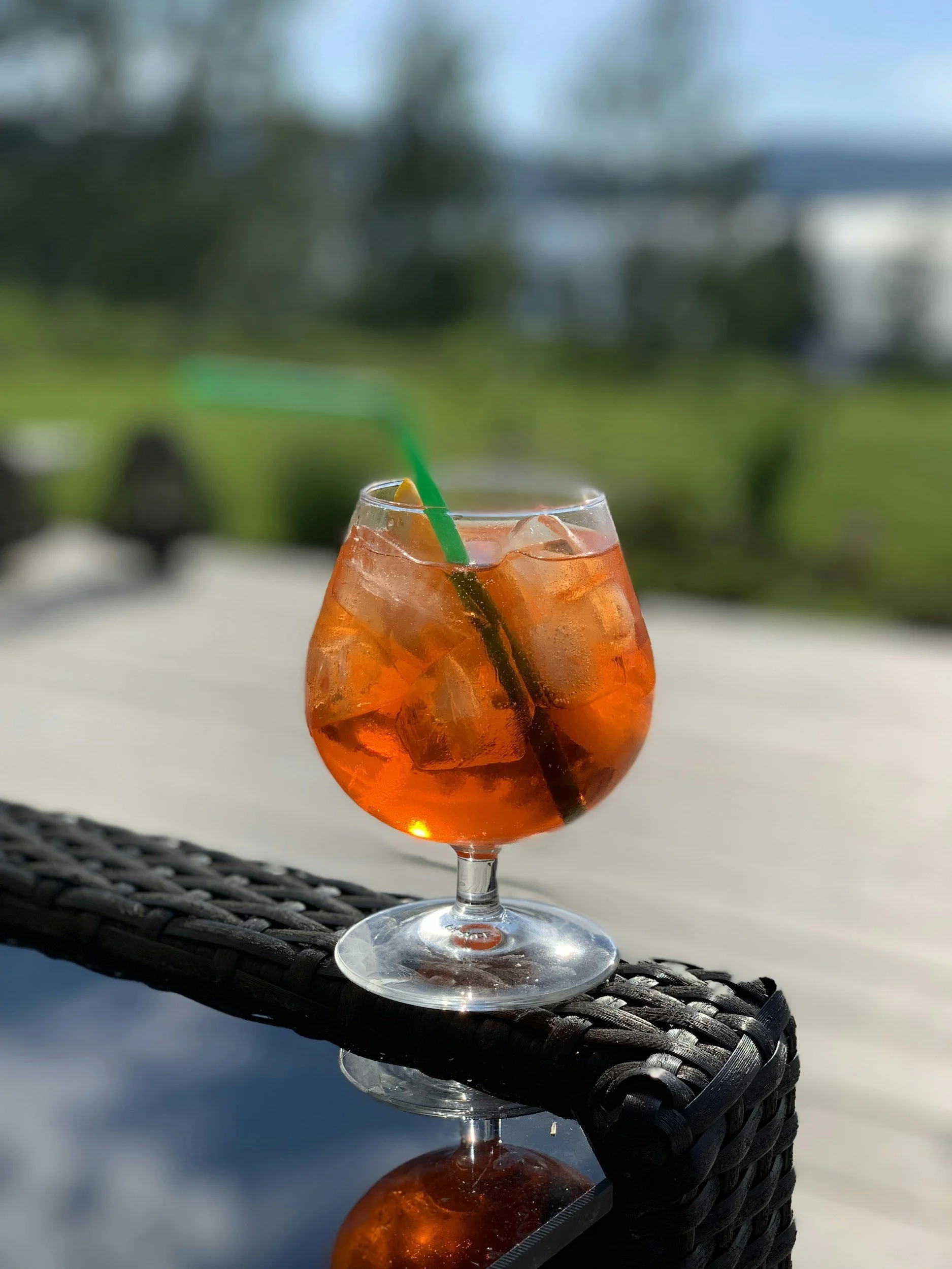 A glass of iced orange-colored drink with a green straw sitting on a wicker table outdoors, with a blurred background of green grass and trees.