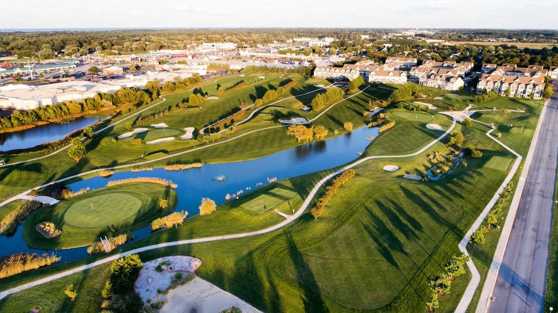 Aerial view of American Classic golf course with water hazards, trees, and pathways, next to a neighborhood with townhouses and commercial area in the background, during daytime.