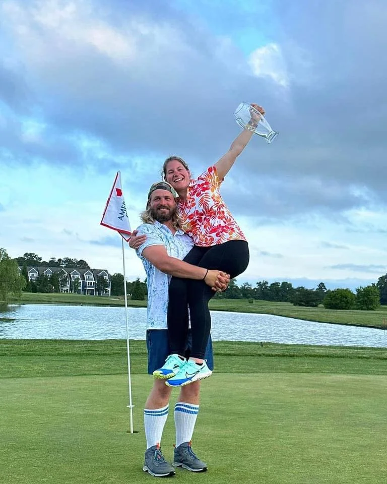 A man and a woman celebrating on a golf course. The man is holding the woman in his arms, she is smiling and holding a trophy, the man is holding a flag. There is a small pond and trees in the background.