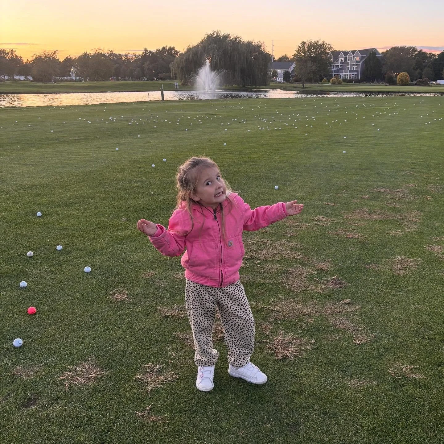 A young girl in a pink jacket and leopard-print pants stands on a golf course with a confused expression, surrounded by scattered golf balls at sunset.