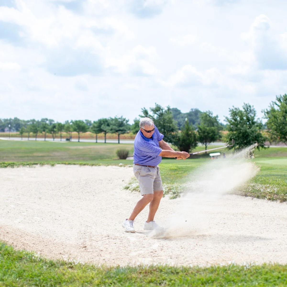 An elderly man playing golf hits a shot out of a sand bunker on a golf course during the day.