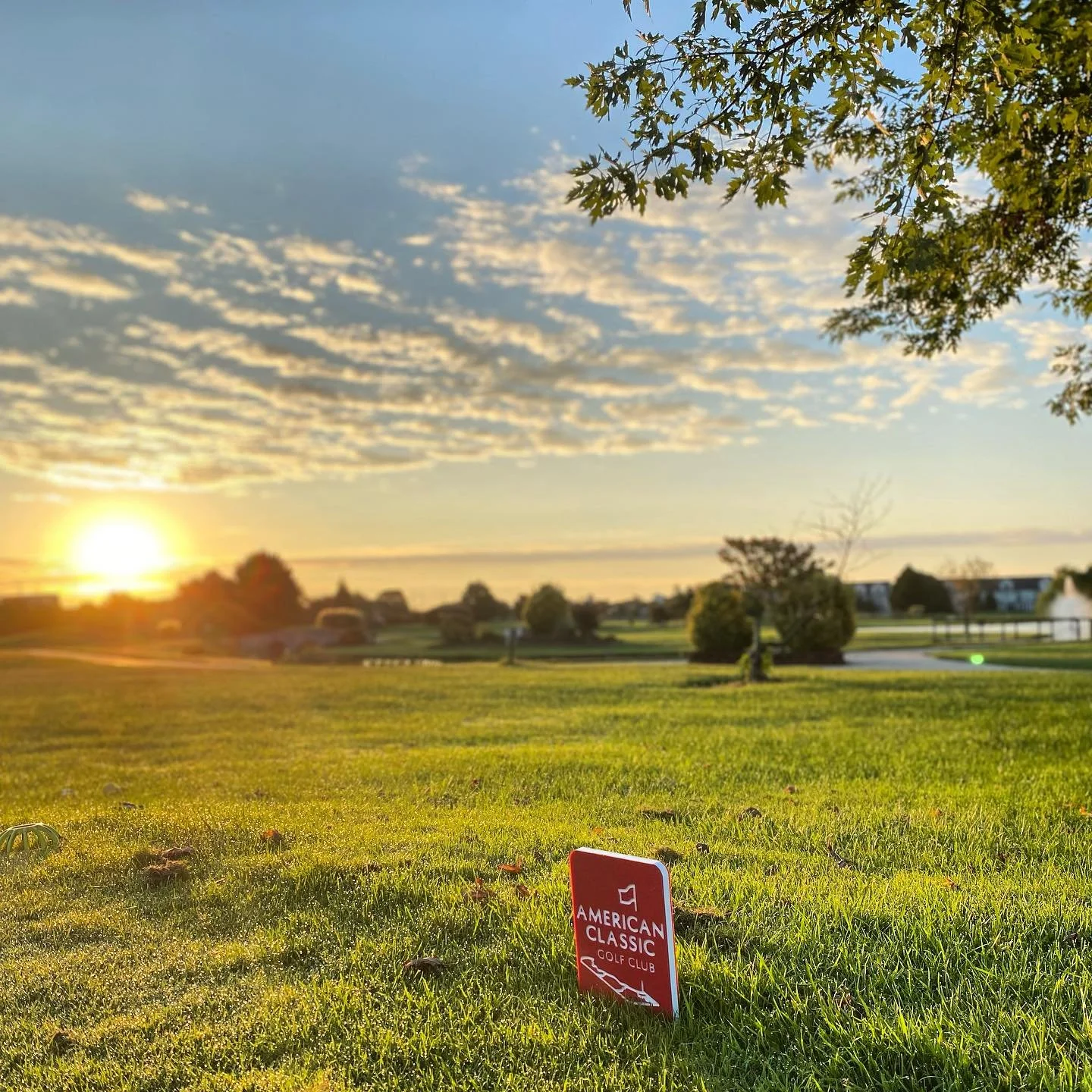 Sunset over a golf course with a green lawn, trees, and a red sign reading 'American Classic Golf Club' in the foreground.