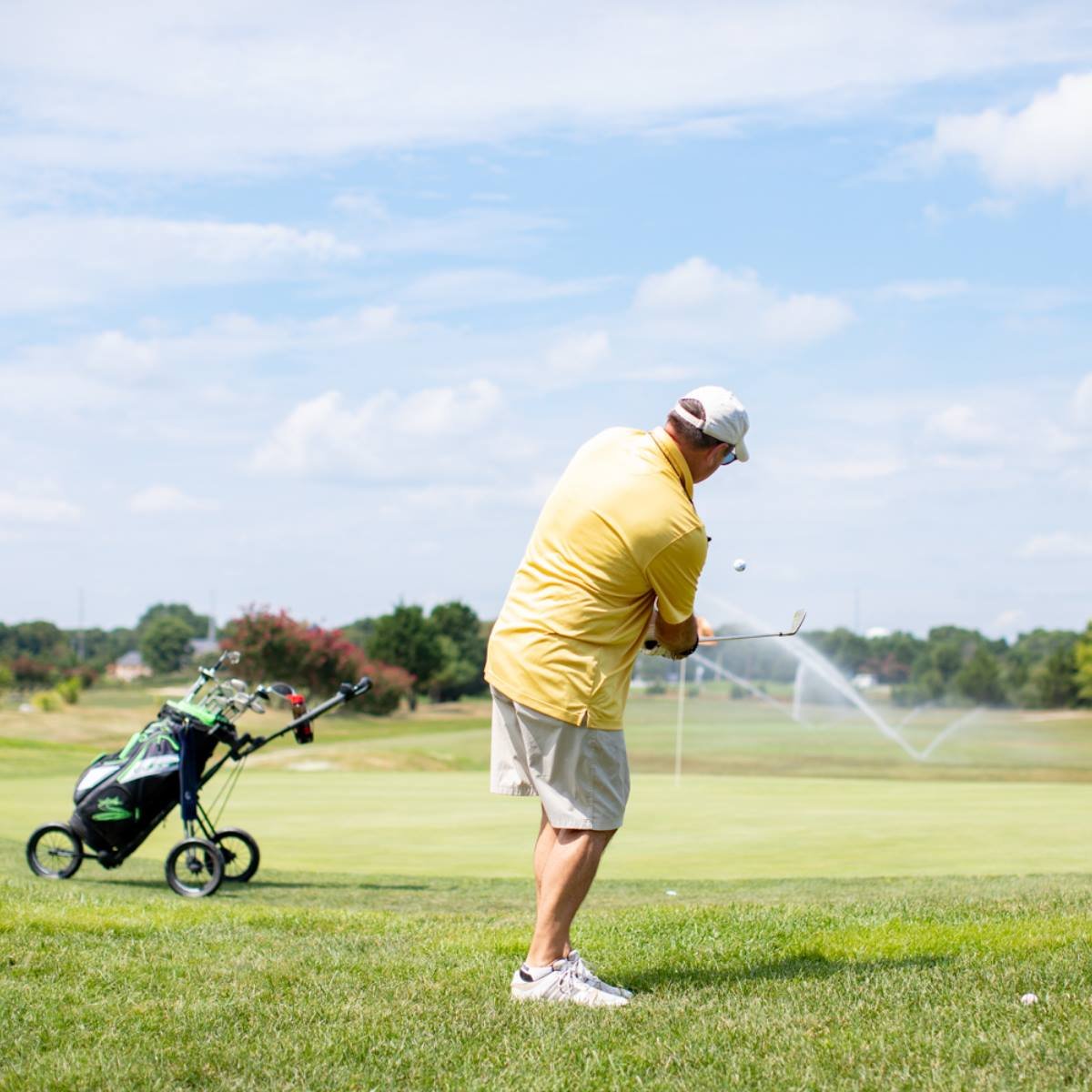 Man playing golf on a sunny day, swinging his golf club with a golf bag and cart nearby.