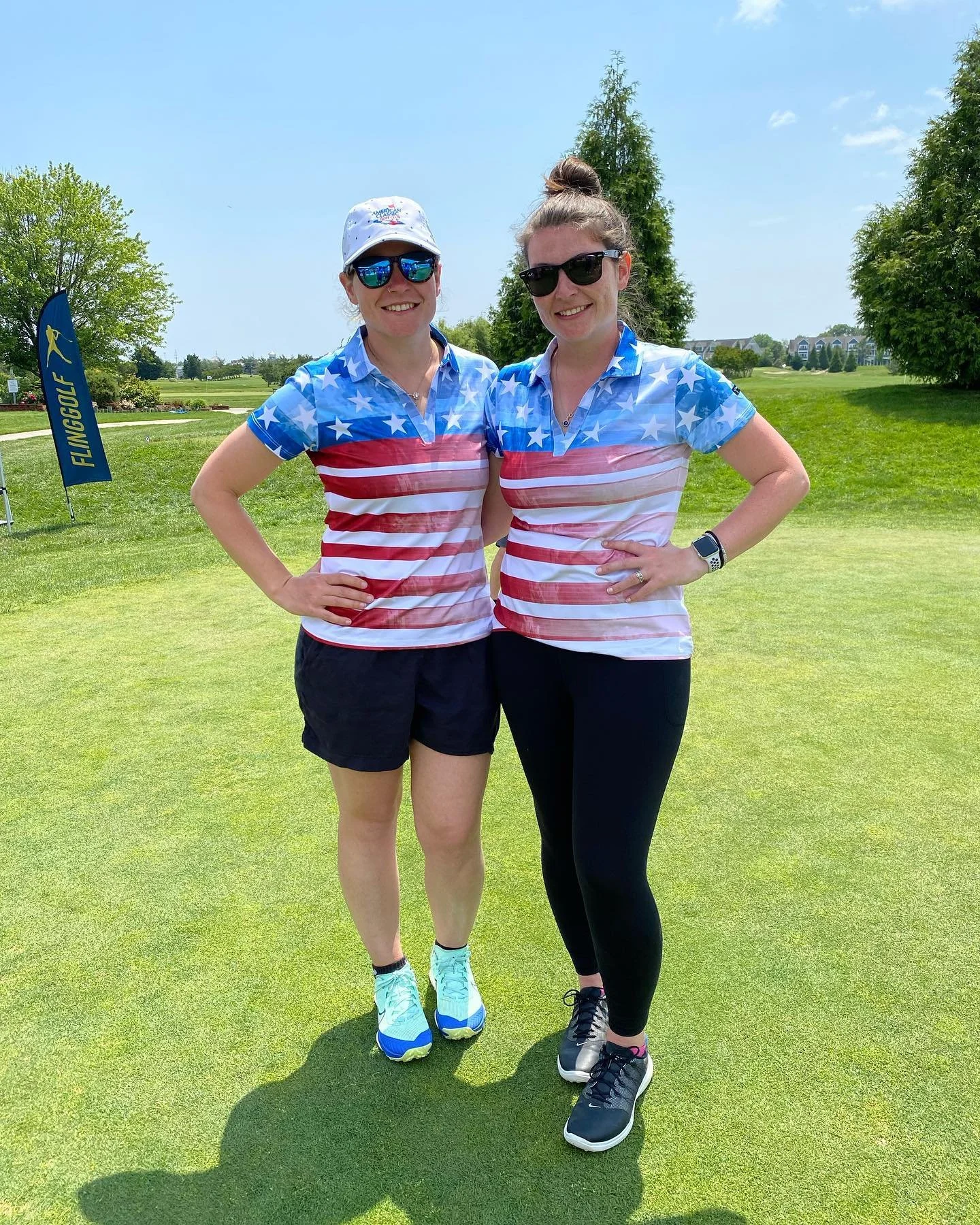 Two women standing on a golf course wearing patriotic shirts with stars and stripes, sunglasses, and athletic shoes, smiling at the camera.