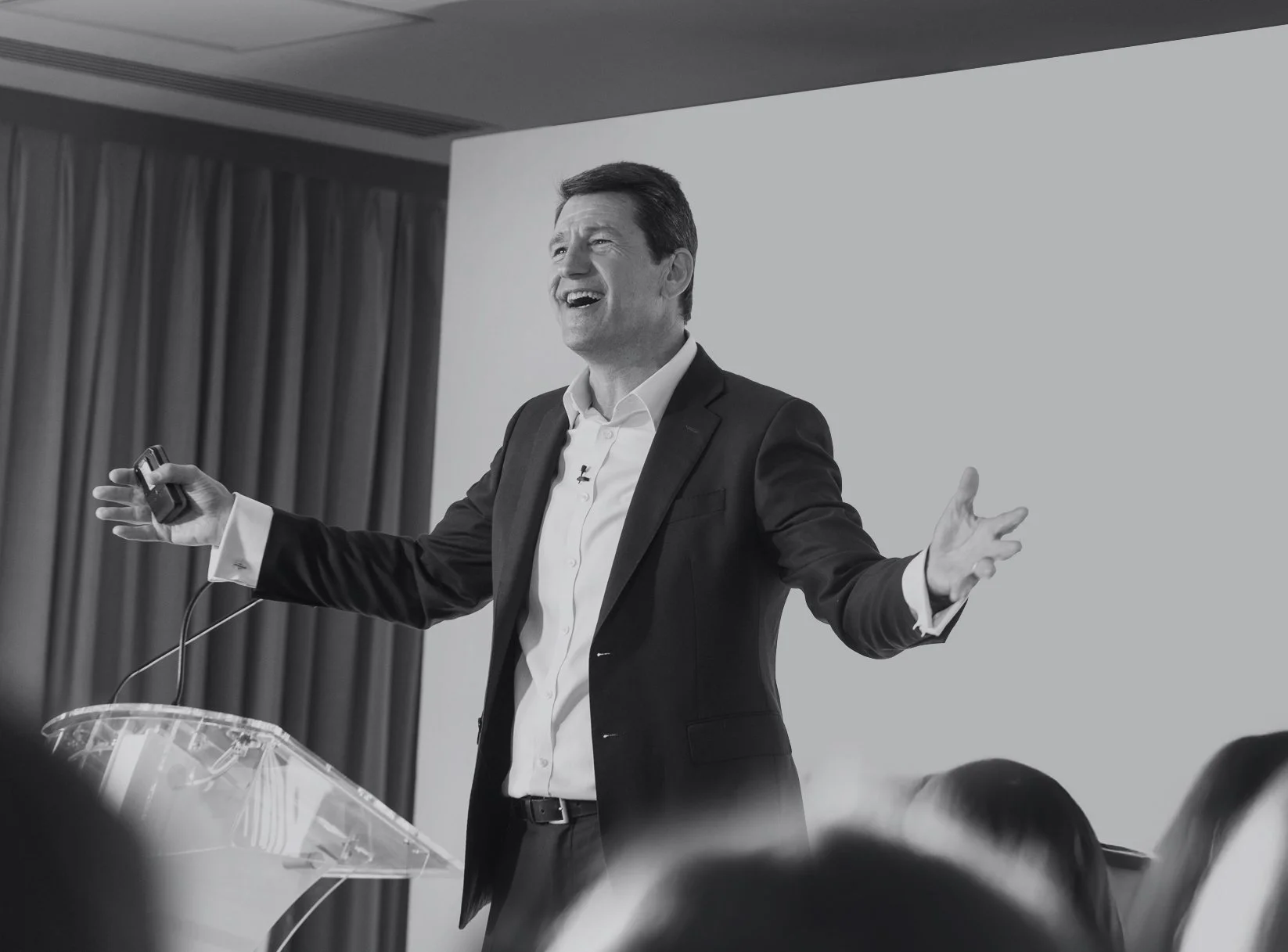 Nick Saunders in a suit giving a presentation, holding a remote in his right hand, smiling, in front of a white screen and dark curtains, during a conference.