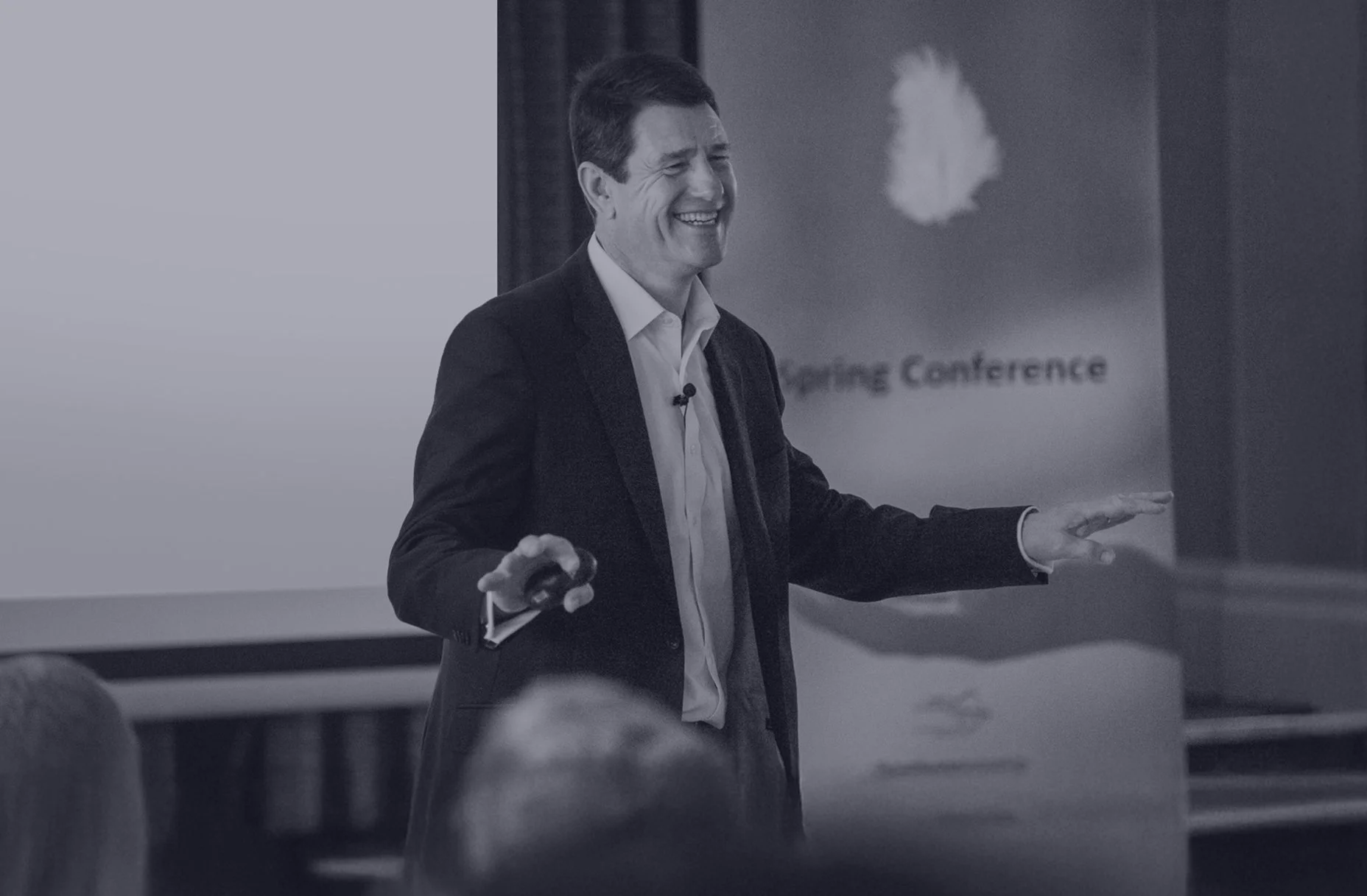Nick Saunders, dressed in a suit, giving a presentation with a smile in a conference room. There is a blurred logo or sign with the words "Spring Conference" in the background.