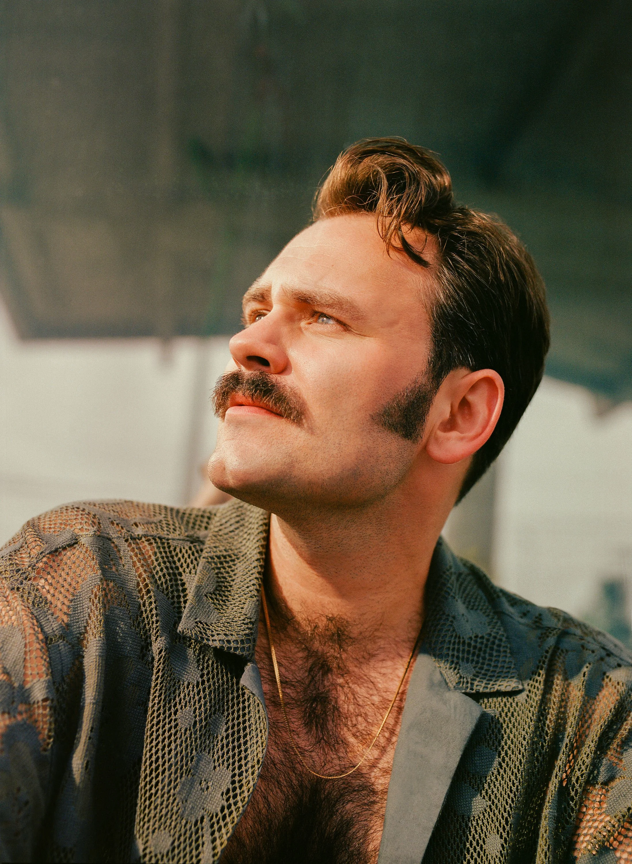 A man with a mustache and sideburns, wearing a patterned shirt and a gold chain, looking to the side with a contemplative expression.