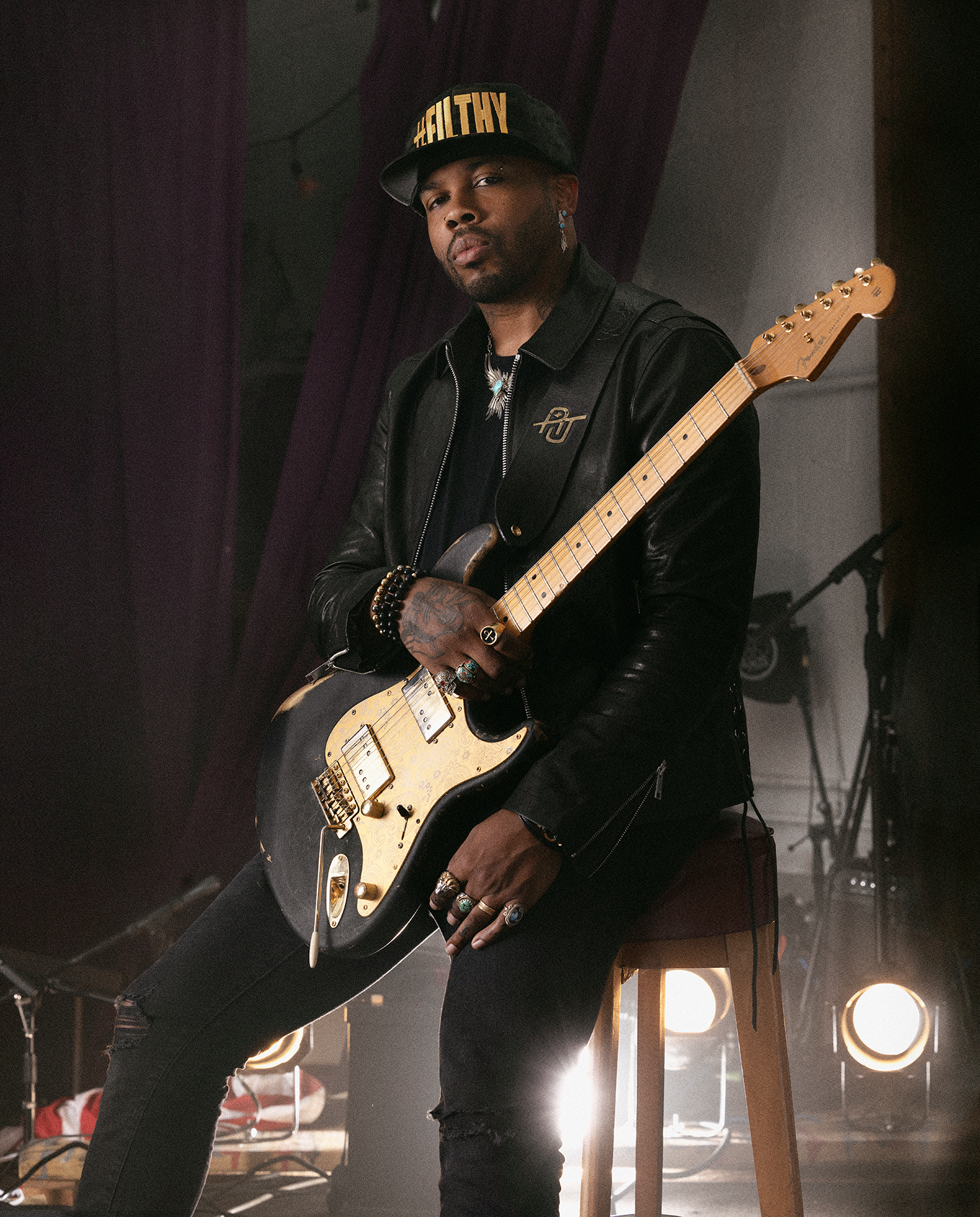 A man sitting on a stool holding an electric guitar in a dimly lit room with stage lights.