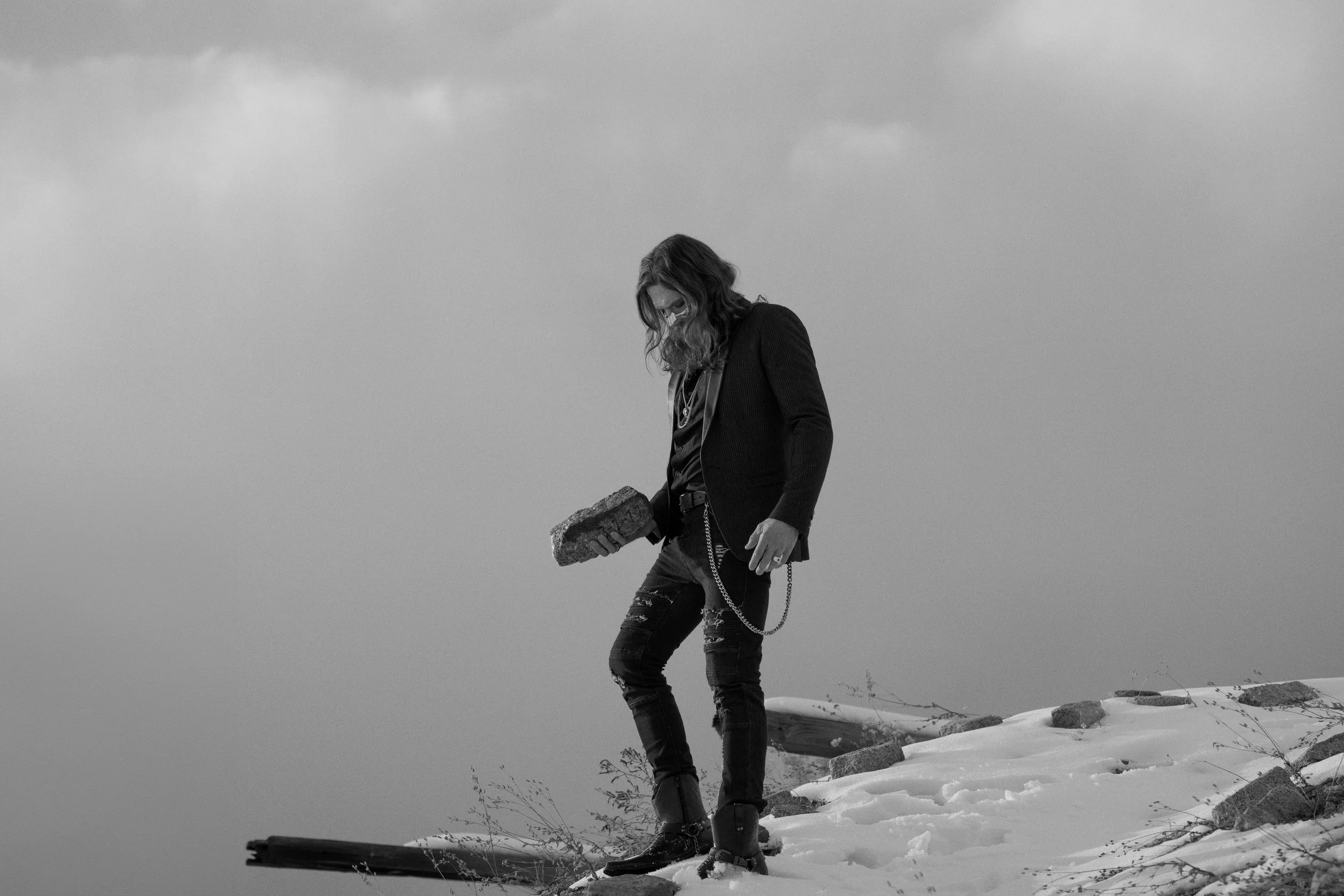 A person with long hair, wearing a dark jacket, ripped jeans, and boots, holding a brick while standing on a snowy hill with rocks and sparse vegetation, under a cloudy sky.