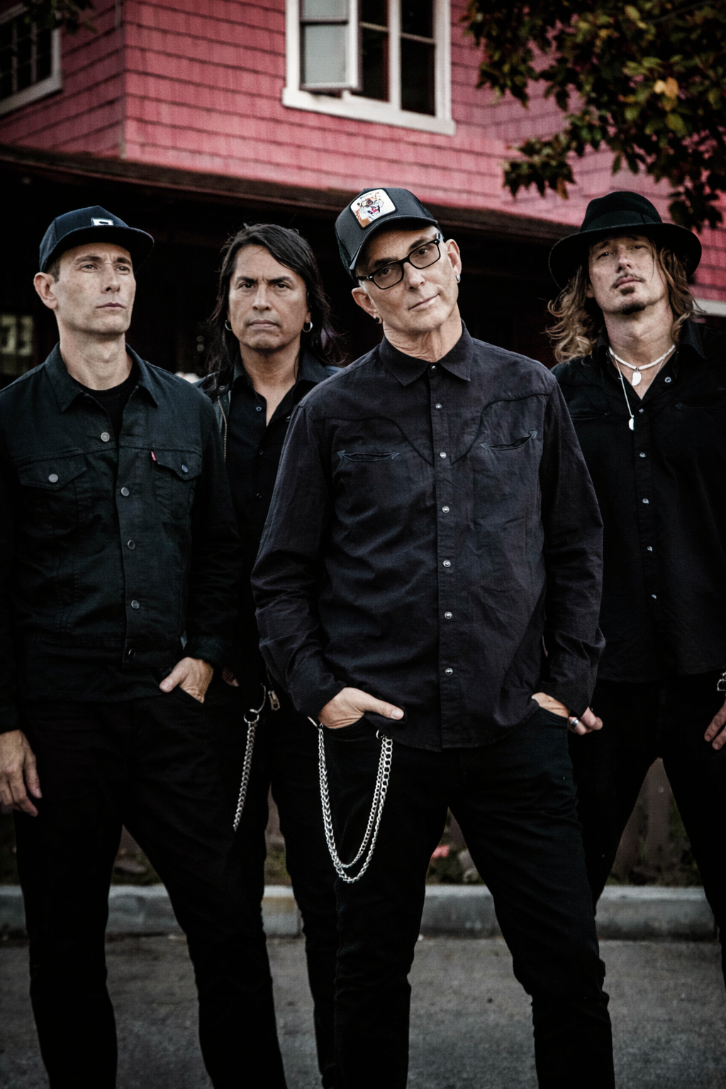 A group of four men standing outdoors in front of a pink house with trees. They are dressed in black clothing, with some wearing hats and chains, and looking serious.