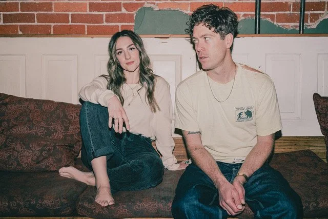 A woman with long hair sitting on a couch next to a man with curly hair, both in casual clothing, in a room with a brick wall in the background.