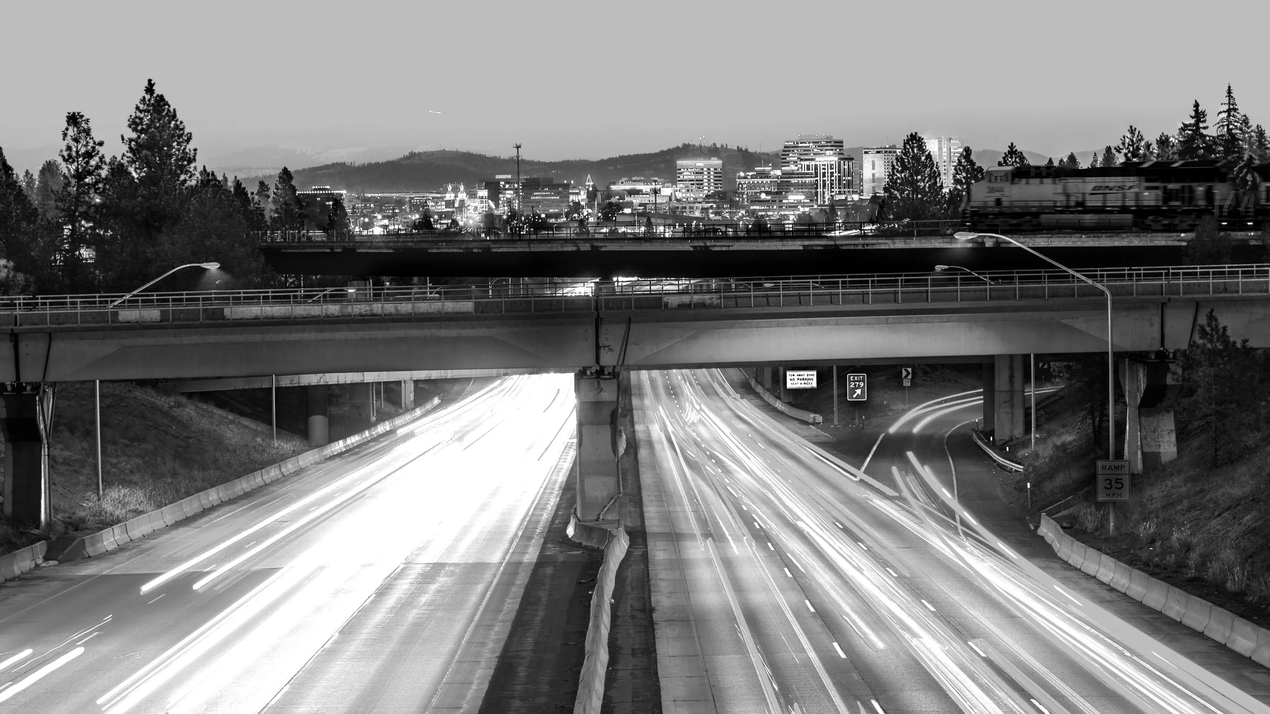 Nighttime view of a busy highway with light trail streaks from passing vehicles, overpasses, city skyline with illuminated buildings, and a train on an elevated track, all in black and white.