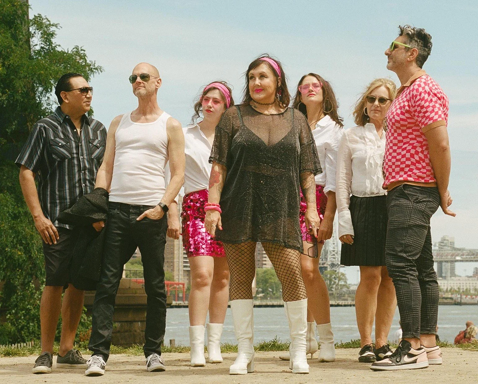 Group of eight people standing outdoors near a body of water, dressed in colorful, retro-inspired clothing and sunglasses.