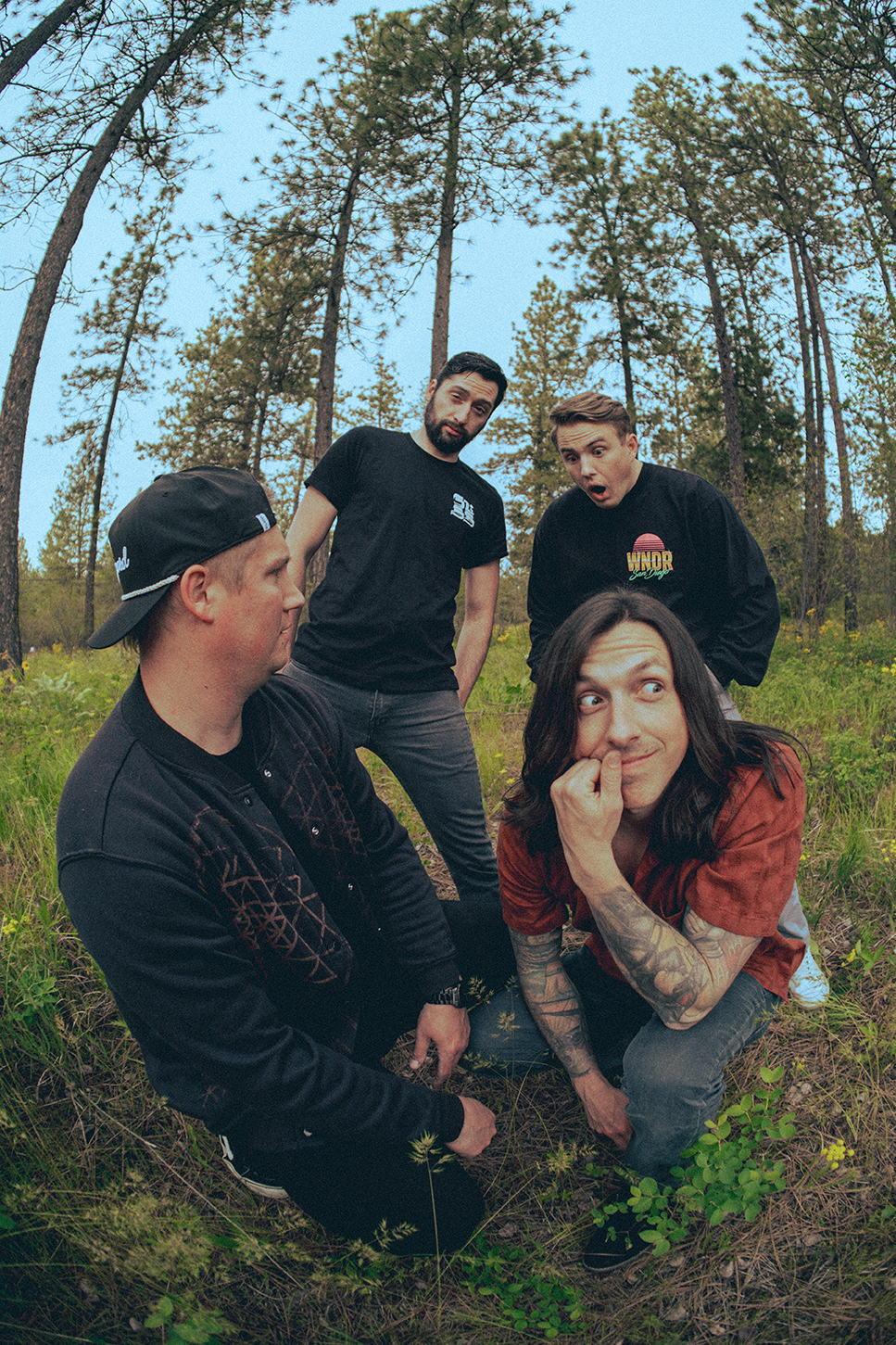 Five young men in casual clothing, some with tattoos and one wearing a backwards baseball cap, are outdoors in a forest clearing, posing playfully for the camera during daytime.