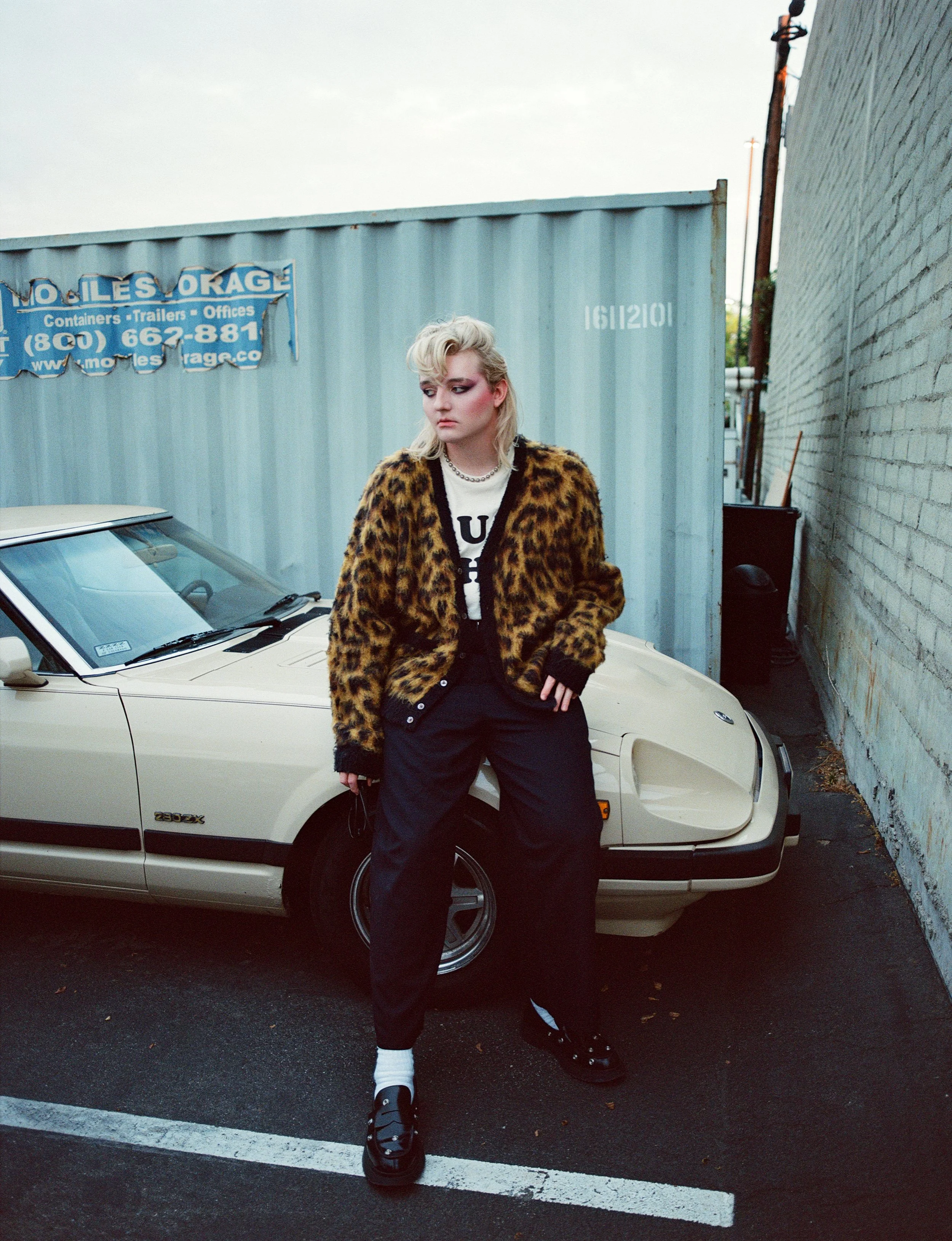 Young woman with blonde hair wearing a leopard print jacket, black pants, and sneakers, standing in front of a vintage cream-colored car with a blue shipping container and brick wall in the background.