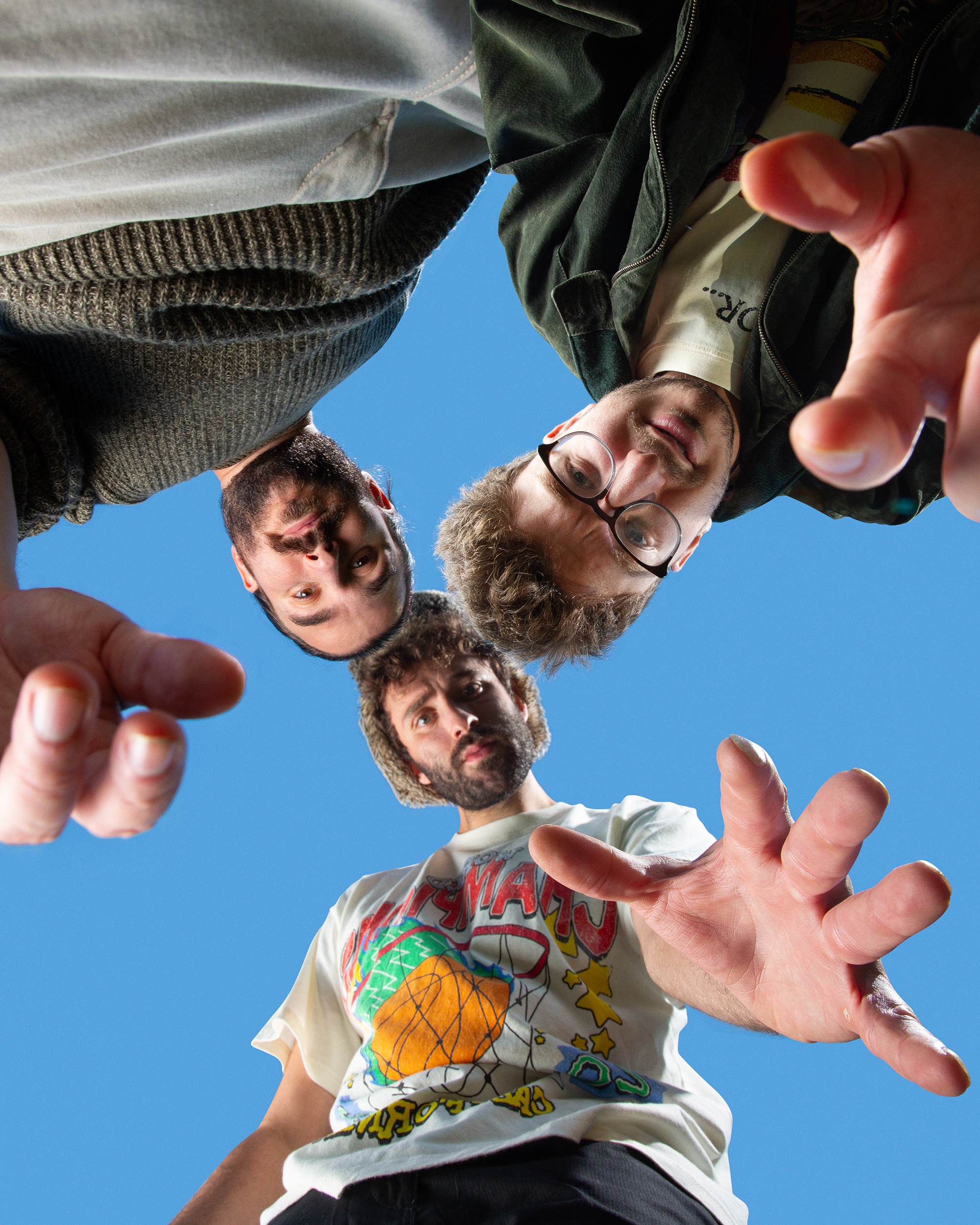 Three young men looking down at the camera with a blue sky background. They are reaching out toward the camera, with only their heads and upper bodies visible.