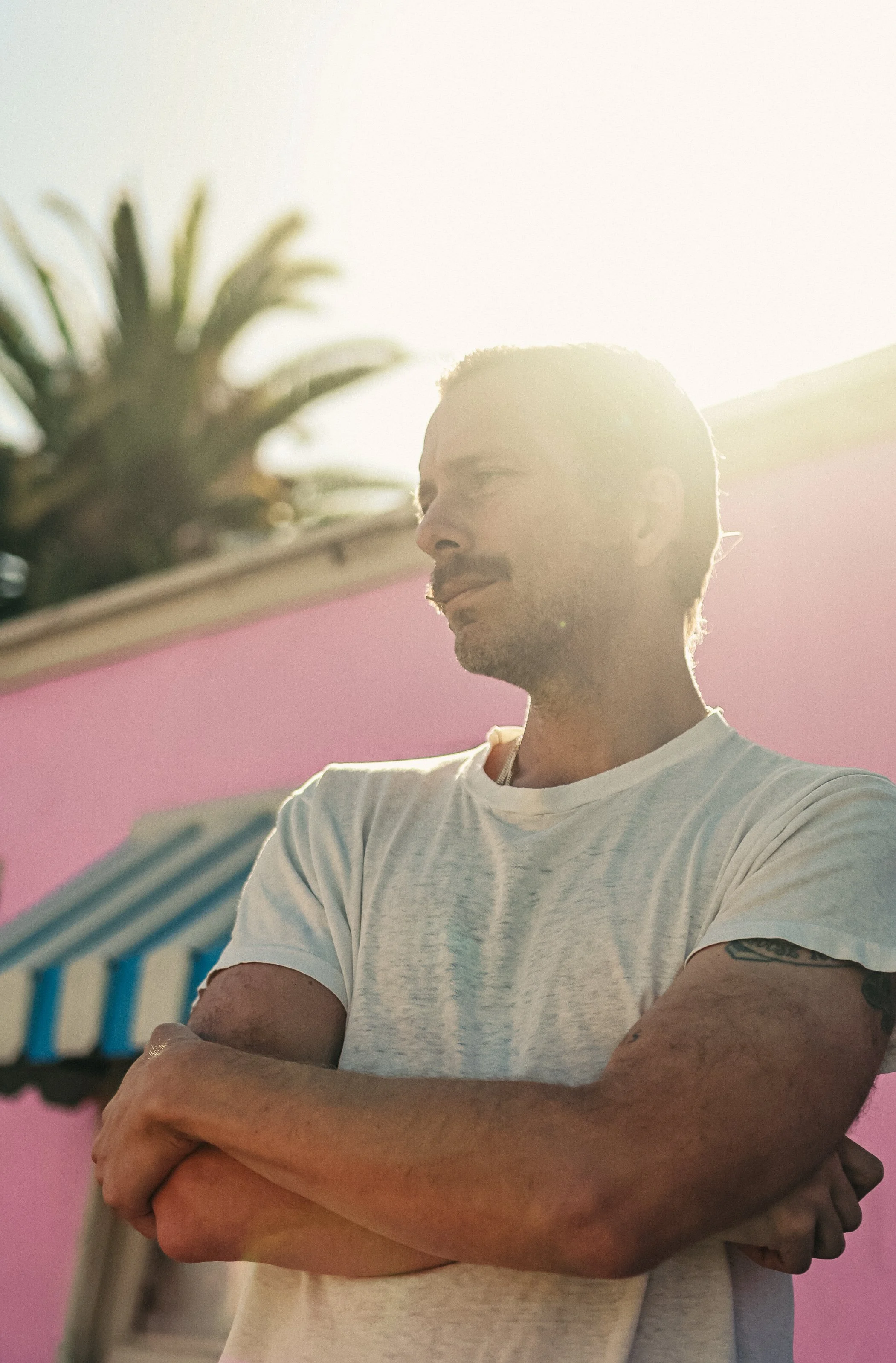 Man with arms crossed standing outdoors in sunlight, wearing a white t-shirt, with a pink building and palm tree in background.