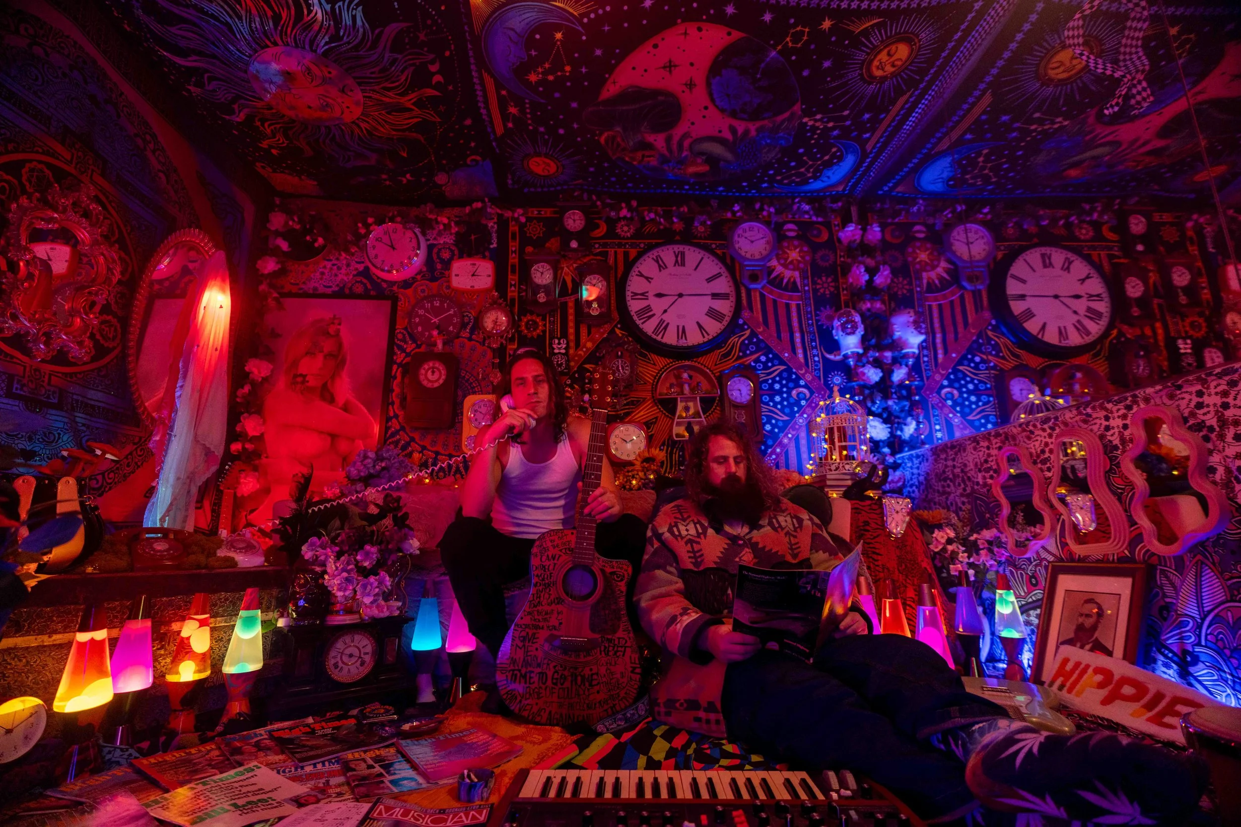 Two men sitting in a psychedelic room filled with clocks, posters, and colorful lights, one holding a guitar and the other reading a magazine.
