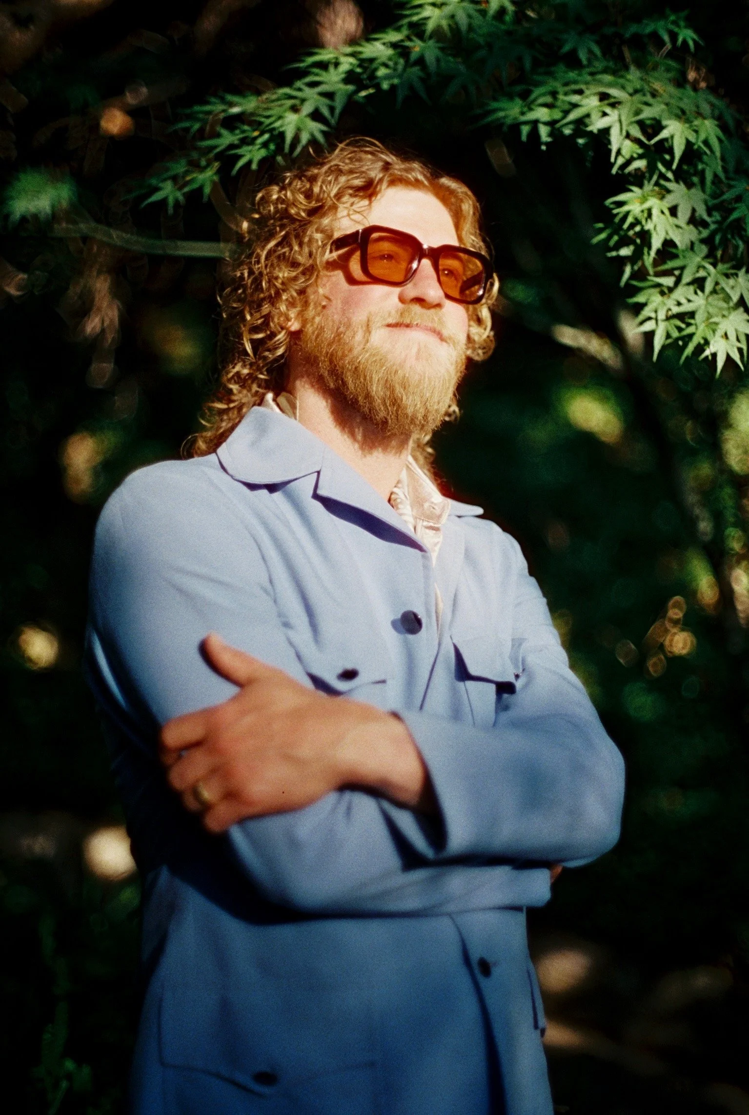 A man with curly hair, a beard, and glasses, wearing a light blue jacket, standing outdoors with arms crossed and green foliage in the background.