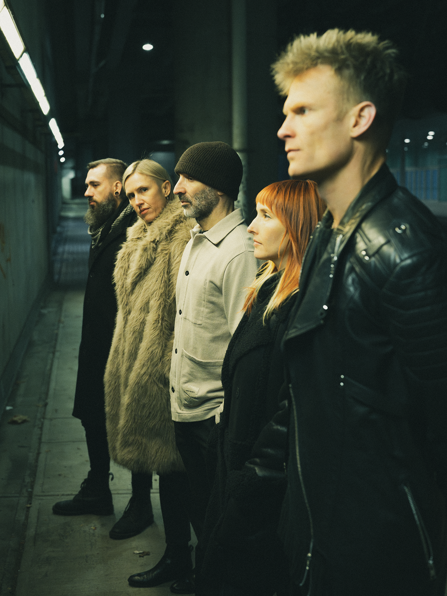 Five people standing in a line in an urban setting at night, wearing dark and stylish clothing, with serious expressions.
