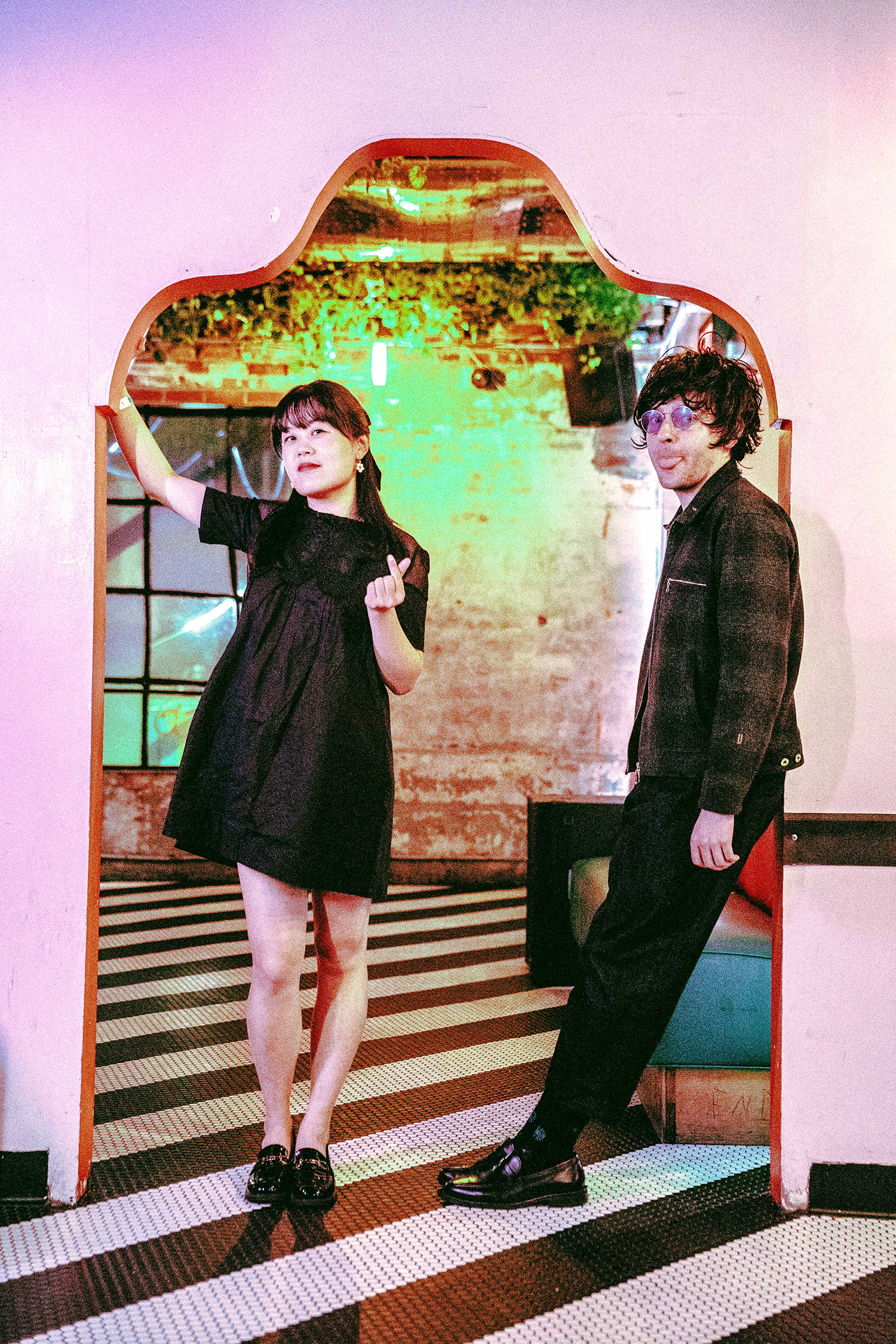 A woman and a man posing in a room with a colorful background, archway, and striped floor. The woman is wearing a black dress and black shoes, while the man is wearing glasses, a black jacket, and dark shoes.