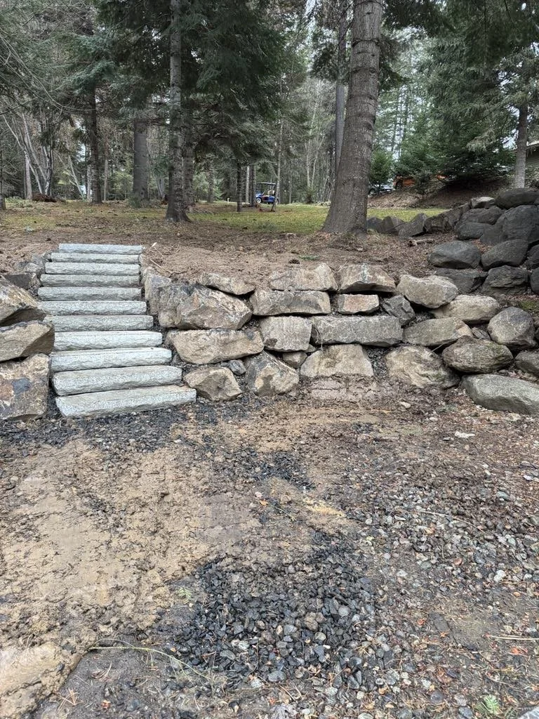 Man wearing green gloves and hat prepping a garden bed with a rake, with gardening equipment including a roller and lawn mower in the background, surrounded by trees and flowers.
