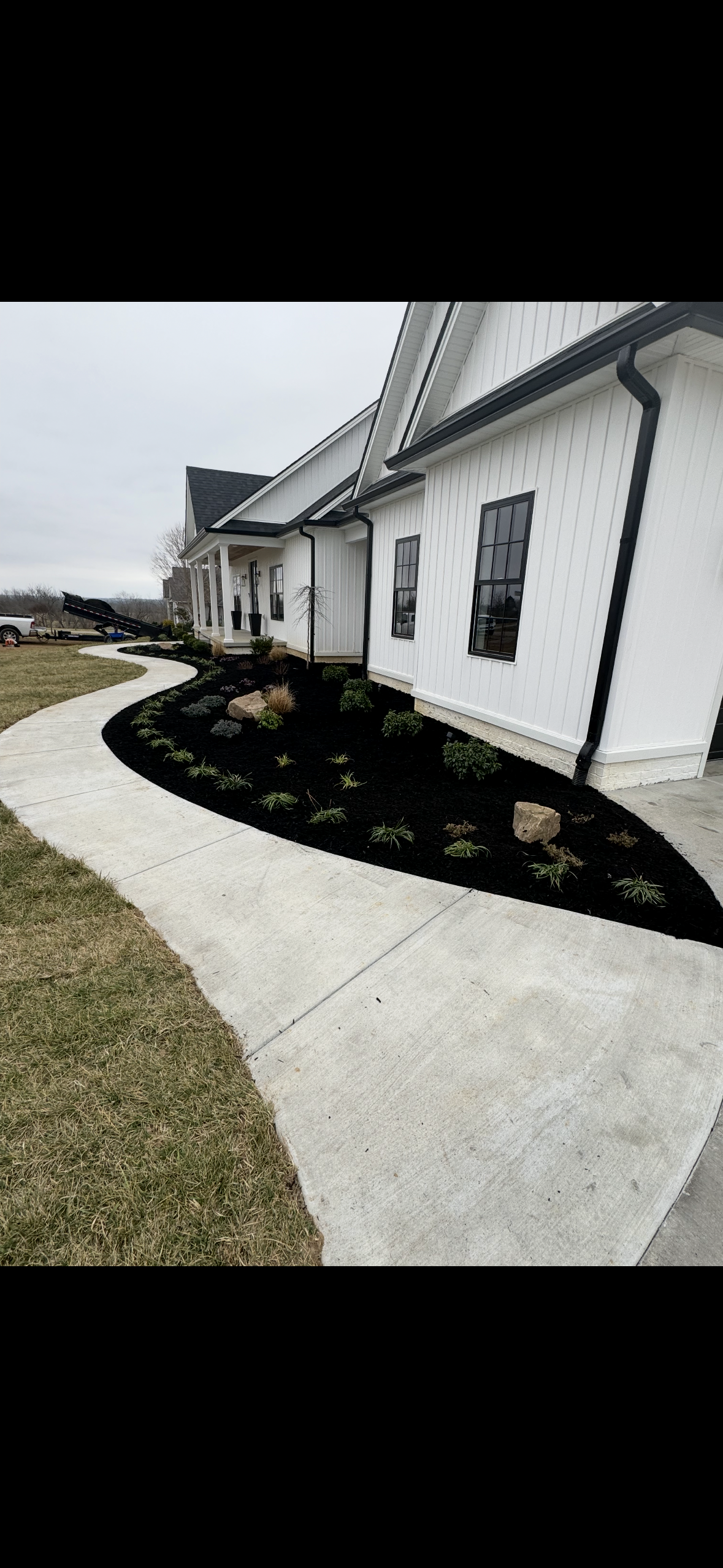 A new white house with black gutters and window frames, landscaped yard with small bushes and rocks, and a curved concrete walkway.