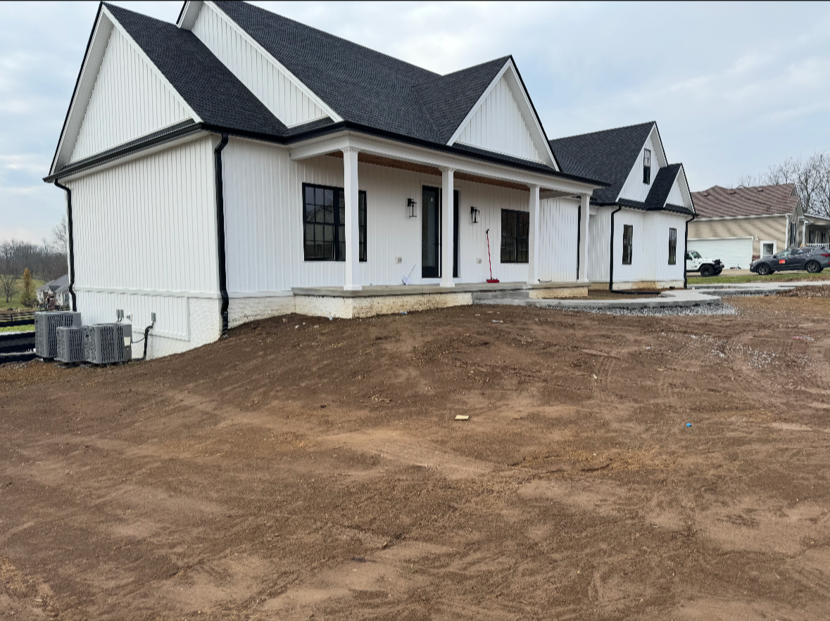 New white house with black roof and front porch, under construction, with disturbed dirt yard and neighboring houses in the background.