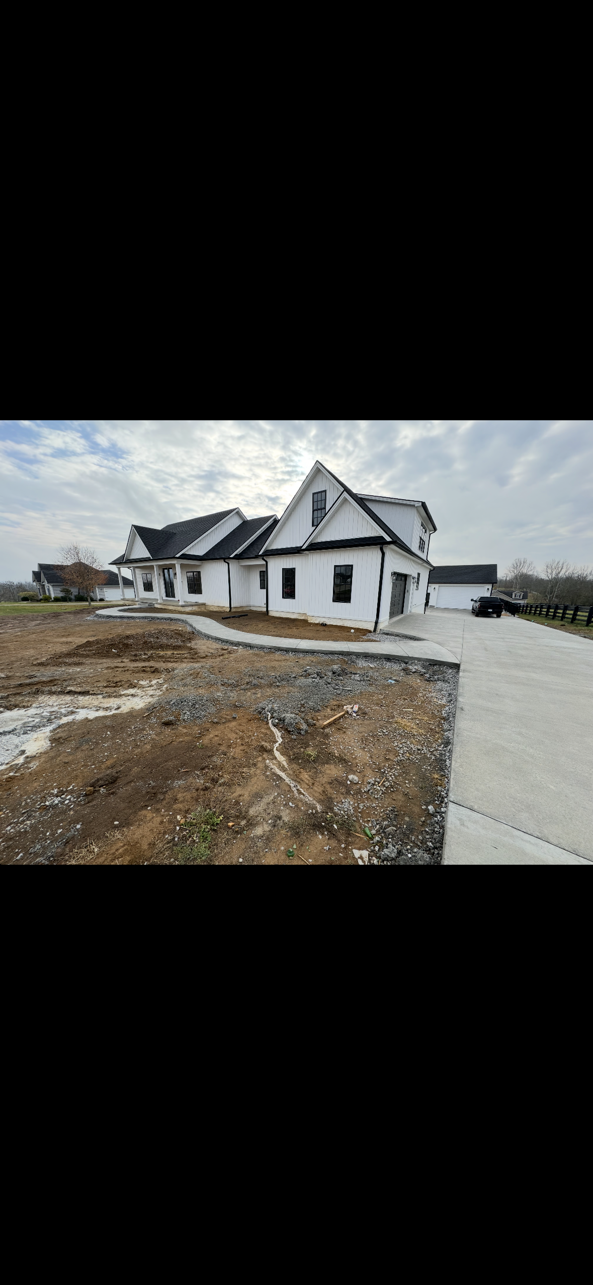Front view of a newly constructed white house with black roofing, surrounded by a freshly paved driveway and a sidewalk, under a partly cloudy sky.