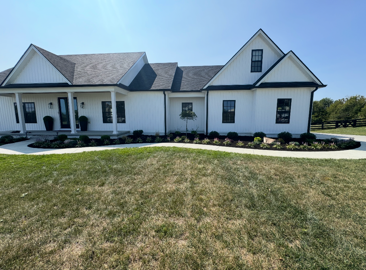 White modern farmhouse-style house with black window frames, front porch, landscaped garden, and lawn under clear blue sky.
