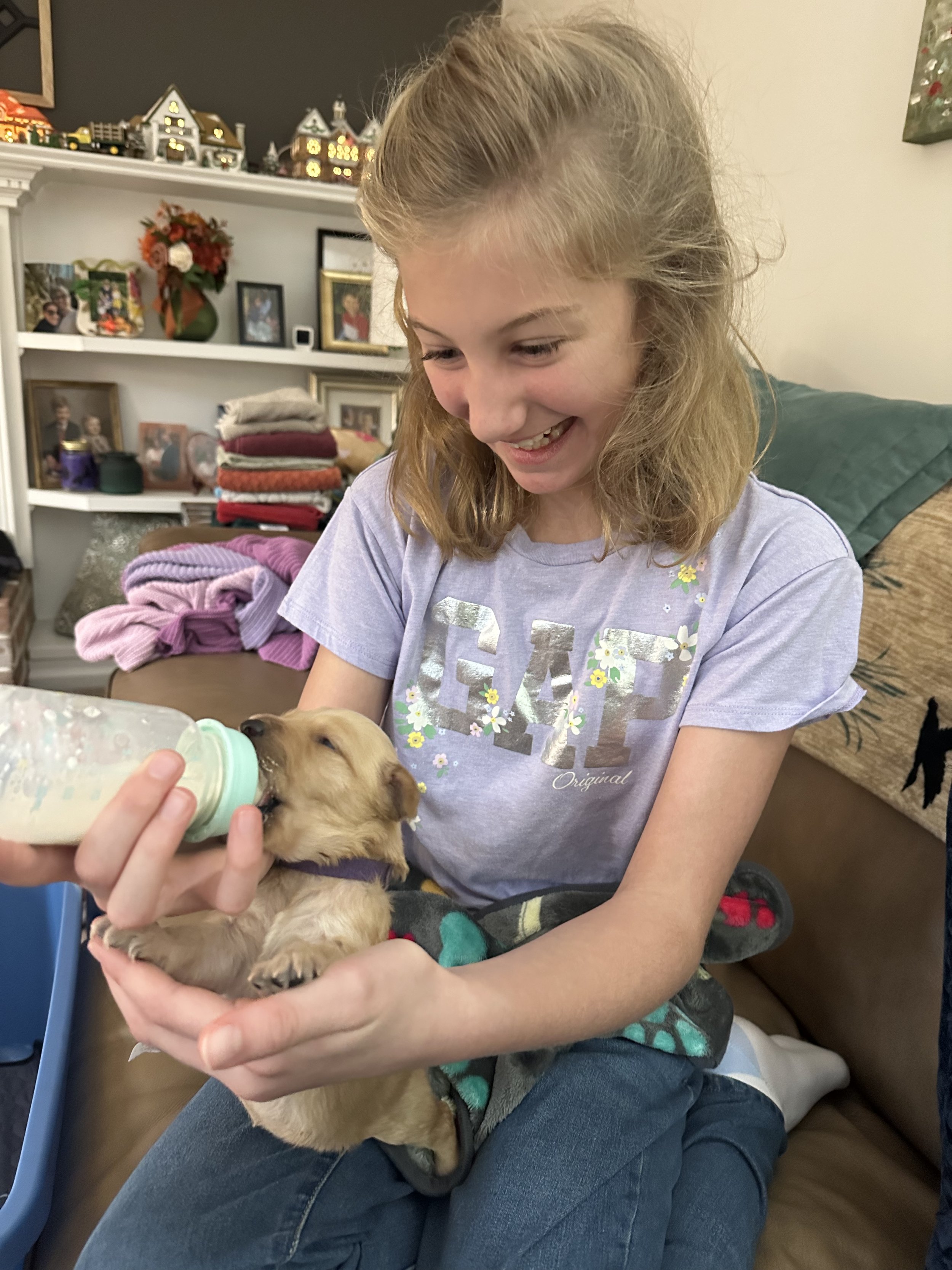 A young girl with blonde hair in a casual T-shirt and jeans is sitting on a couch and bottle-feeding a small puppy. The girl is smiling and looking down at the puppy, which is drinking milk from a baby bottle.