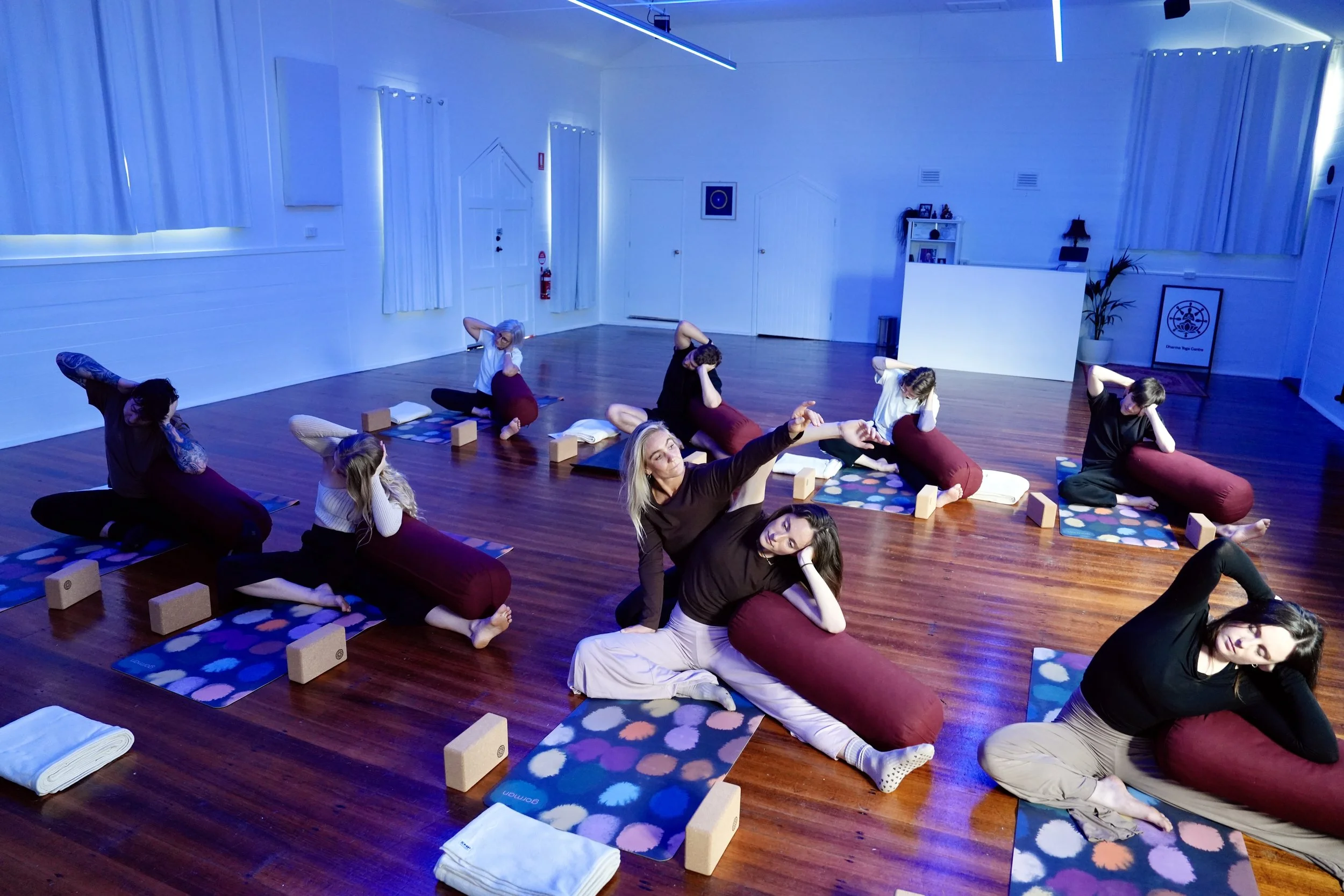 People participating in a yoga class in a room with wooden floors, blue lighting, yoga mats, bolsters, and blocks.