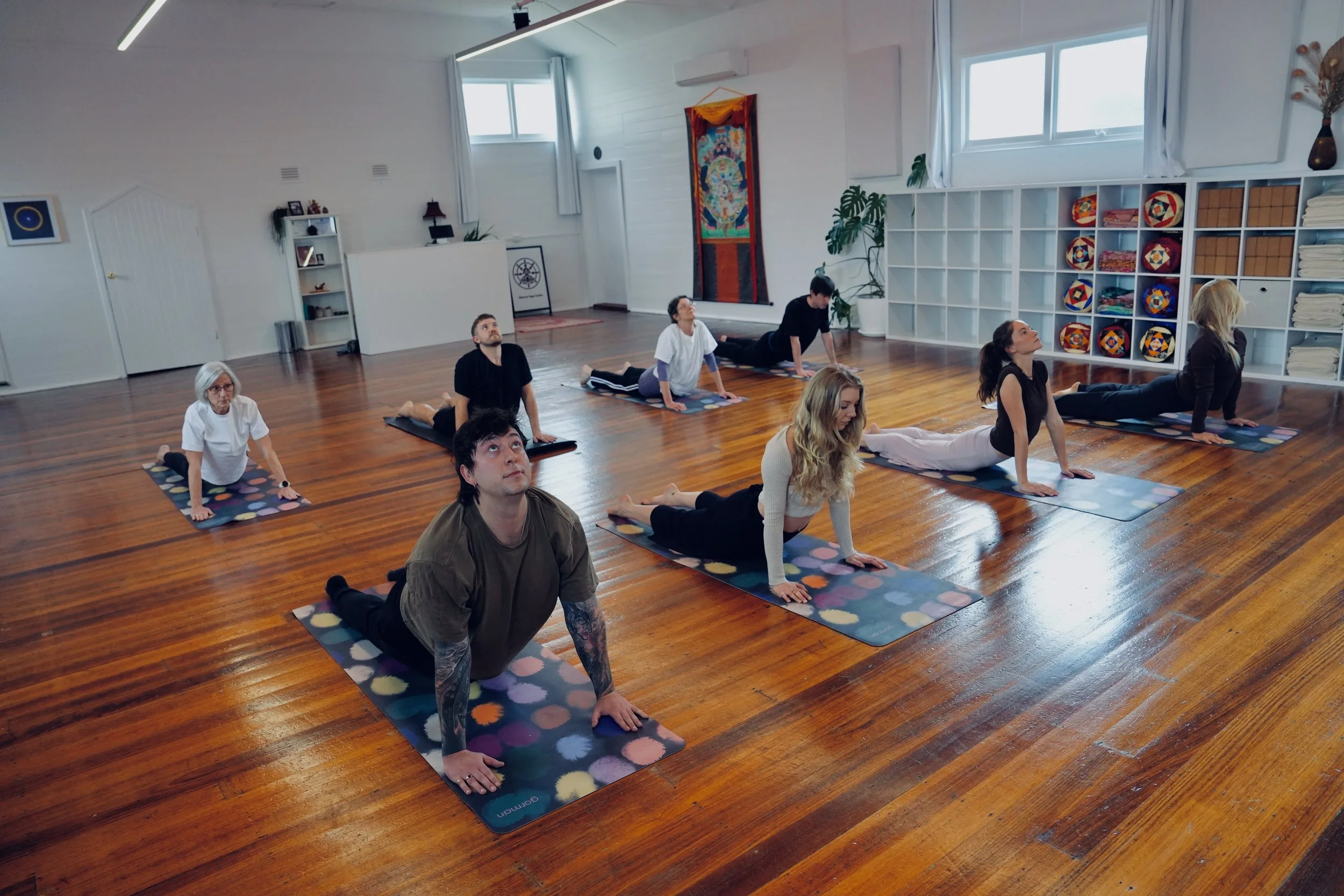 People participating in a yoga class in a bright, modern studio with wooden floors, white walls, large windows, and shelves with colorful yoga props.