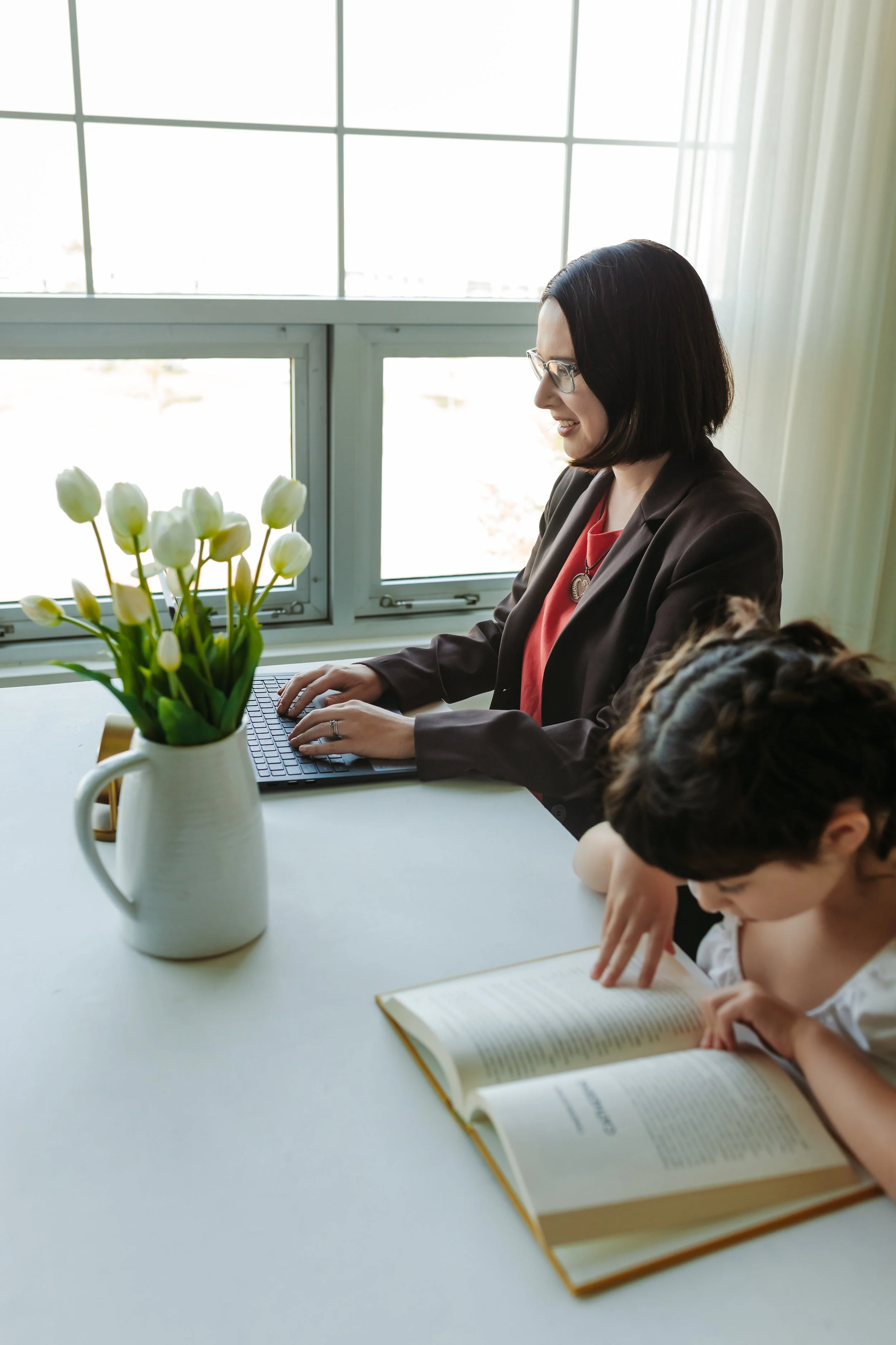 A woman with medium length brown hair, glasses, a brown blazer, working on a laptop with a girl, brown hair and braids, reading a book beside her. On the table is a vase of white tulips, the background is a window.