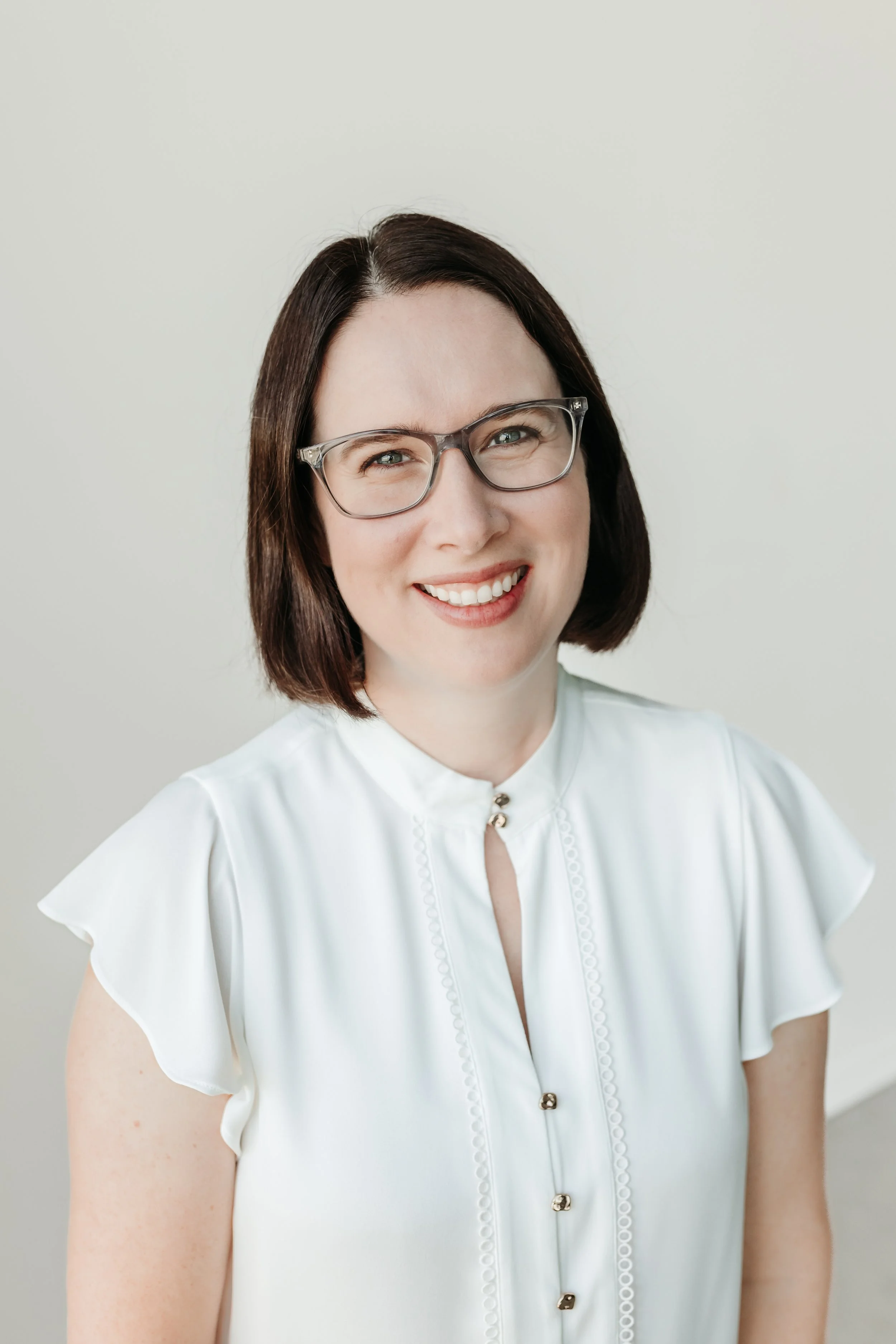 A woman with medium length brown hair and glasses wearing a white blouse smiling at the camera with a neutral background