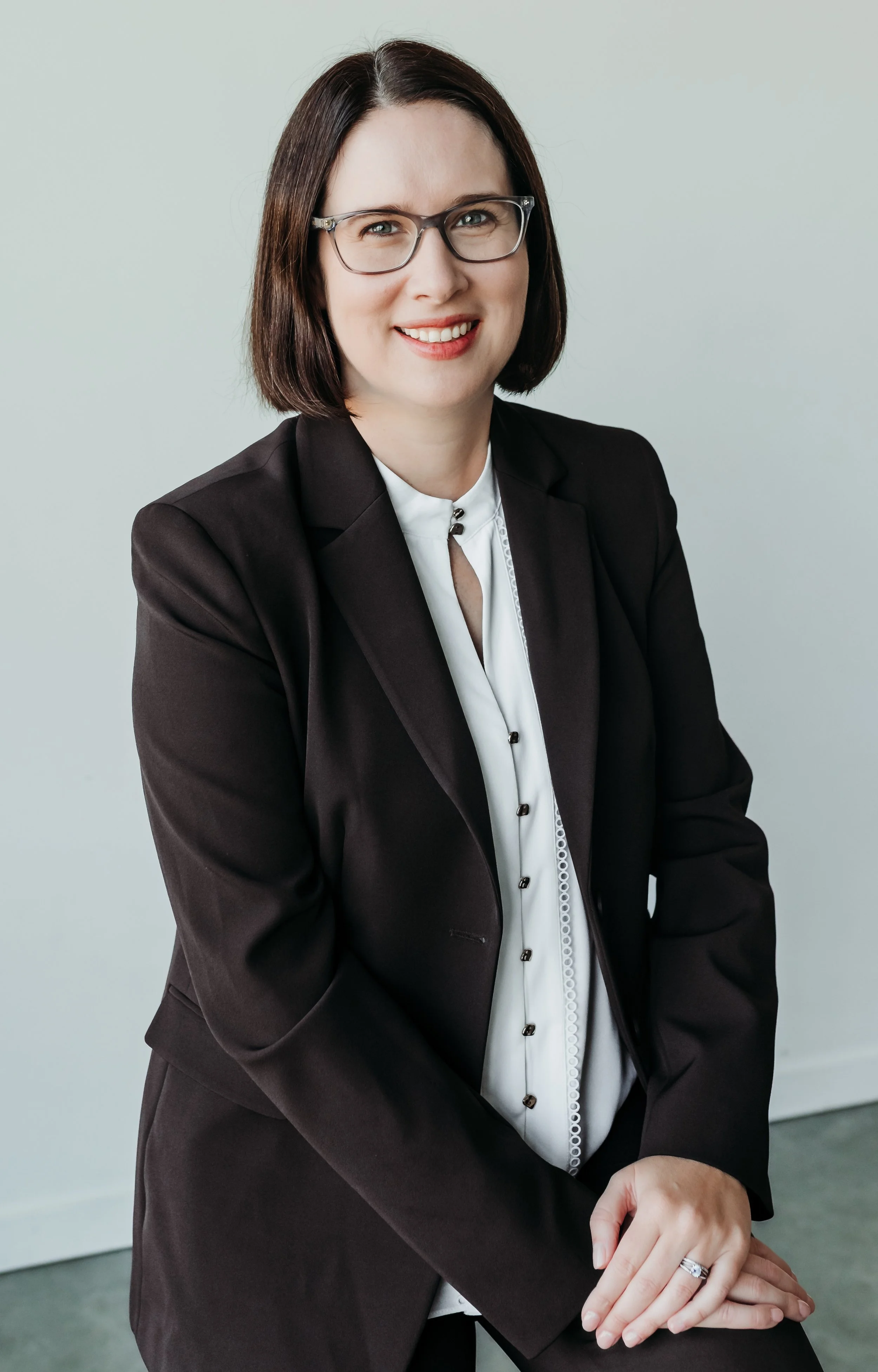 A woman with medium length brown hair and glasses, wearing a brown blazer and white top, smiling with hands clasped on her knee, against a plain grey background.